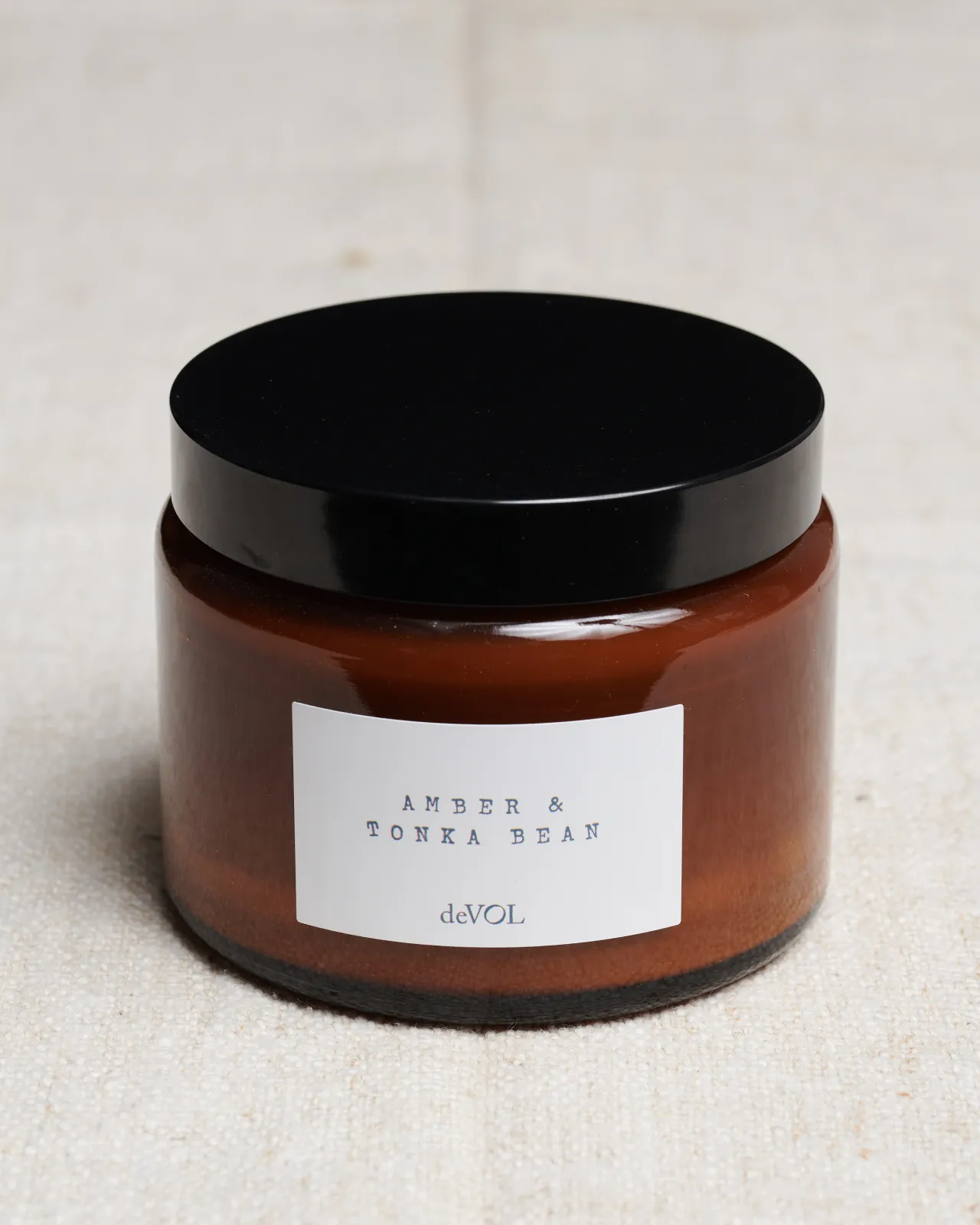 A large scented candle in a brown glass jar with a black lid, the label reads 'Amber & Tonka Bean, deVOL'.