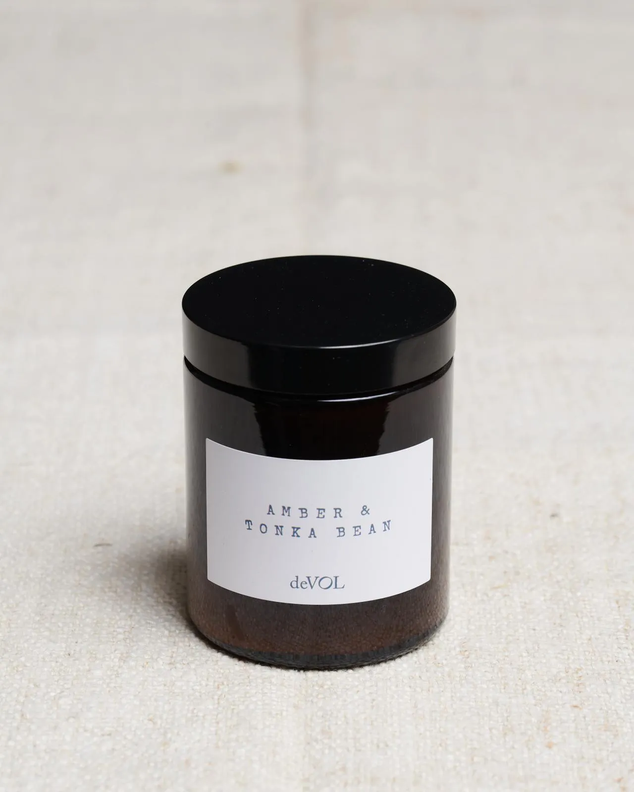 A medium scented candle in a brown glass jar with a black lid, the label reads 'Amber & Tonka Bean, deVOL'.