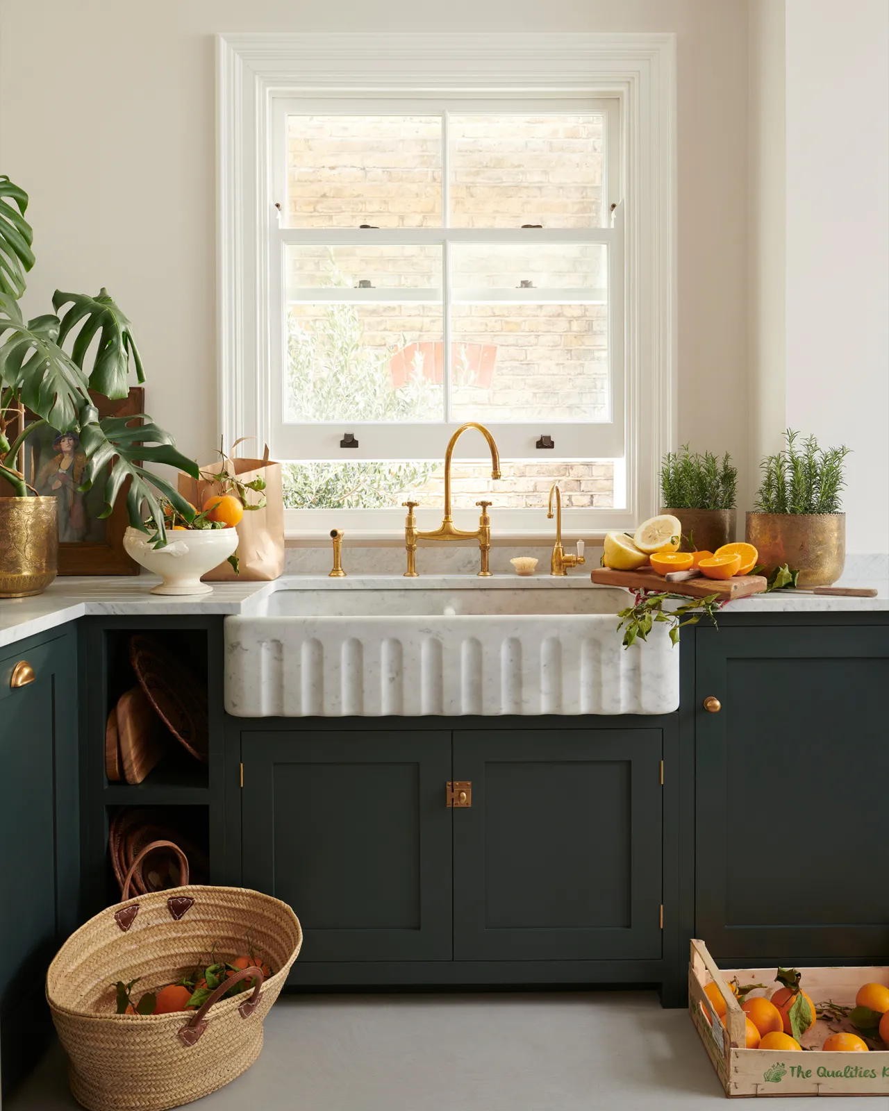 A dark green painted Shaker-style kitchen with light coloured walls, a cement floor, a wide Carrara marble sink with a fluted front, aged brass taps, a monstera plant and baskets of vibrant oranges.