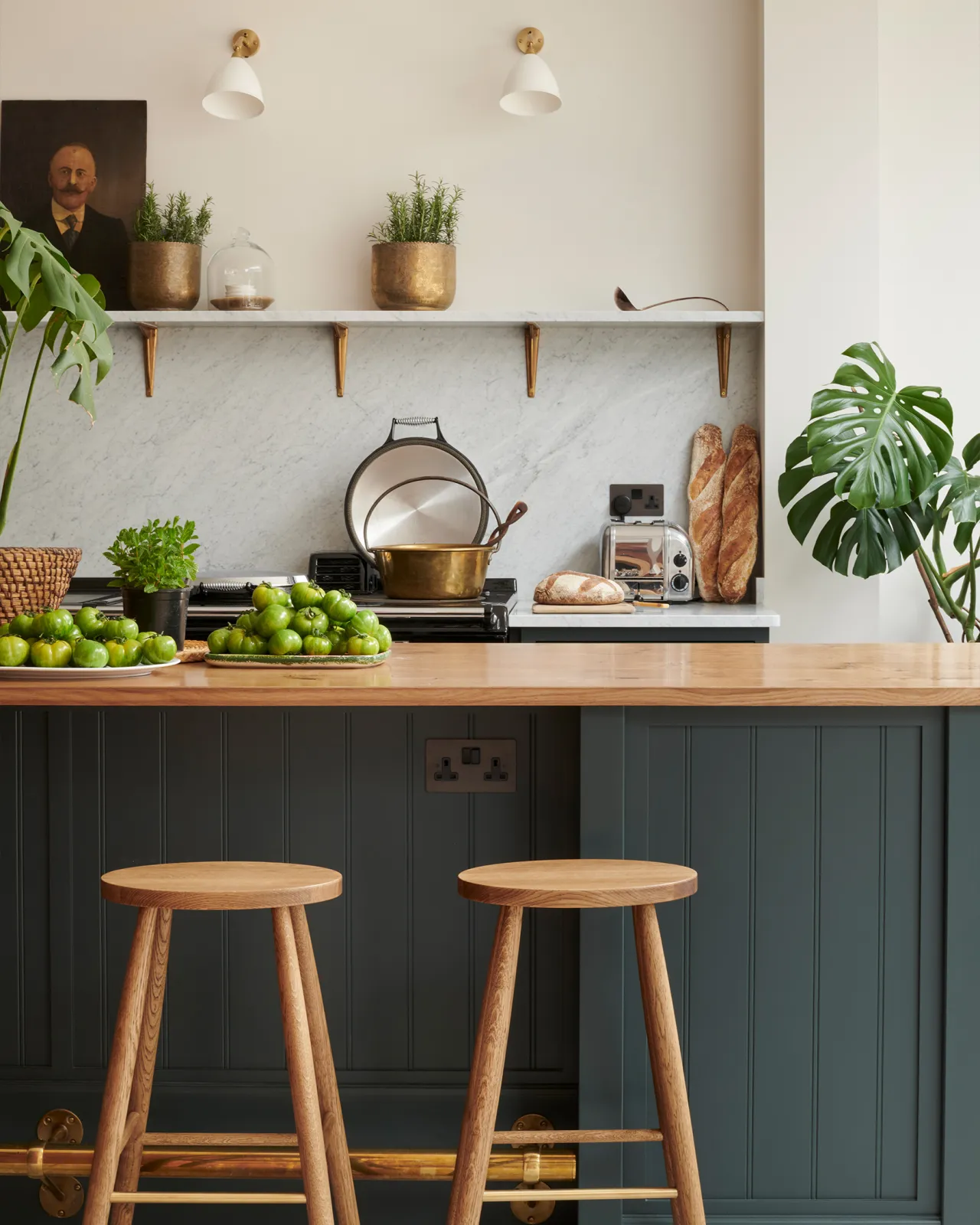 A dark green painted Shaker-style kitchen with a breakfast bar island, simple oak bar stools and a Carrara marble splashback.