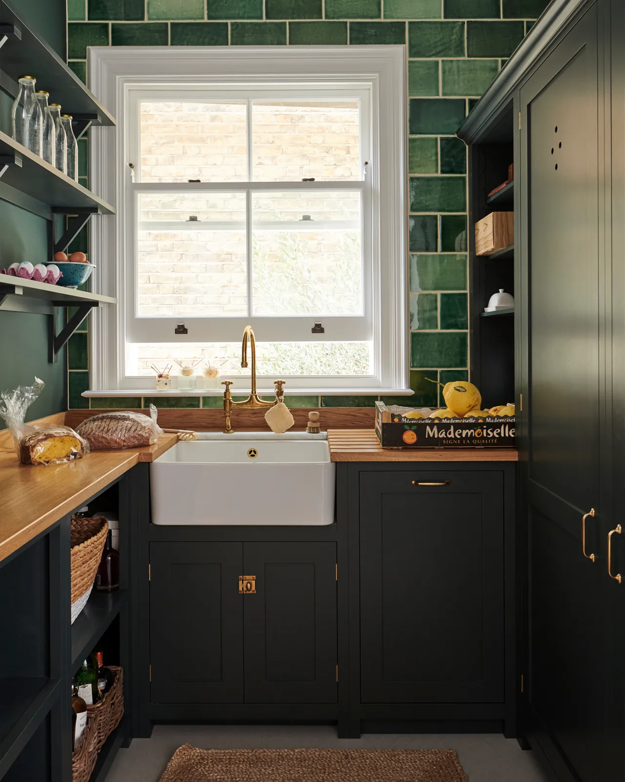 A moody utility kitchen with dark green painted Shaker-style cupboards, a small Belfast sink below a large sash window, hardwood worktops and a wall of glossy emerald green tiles.