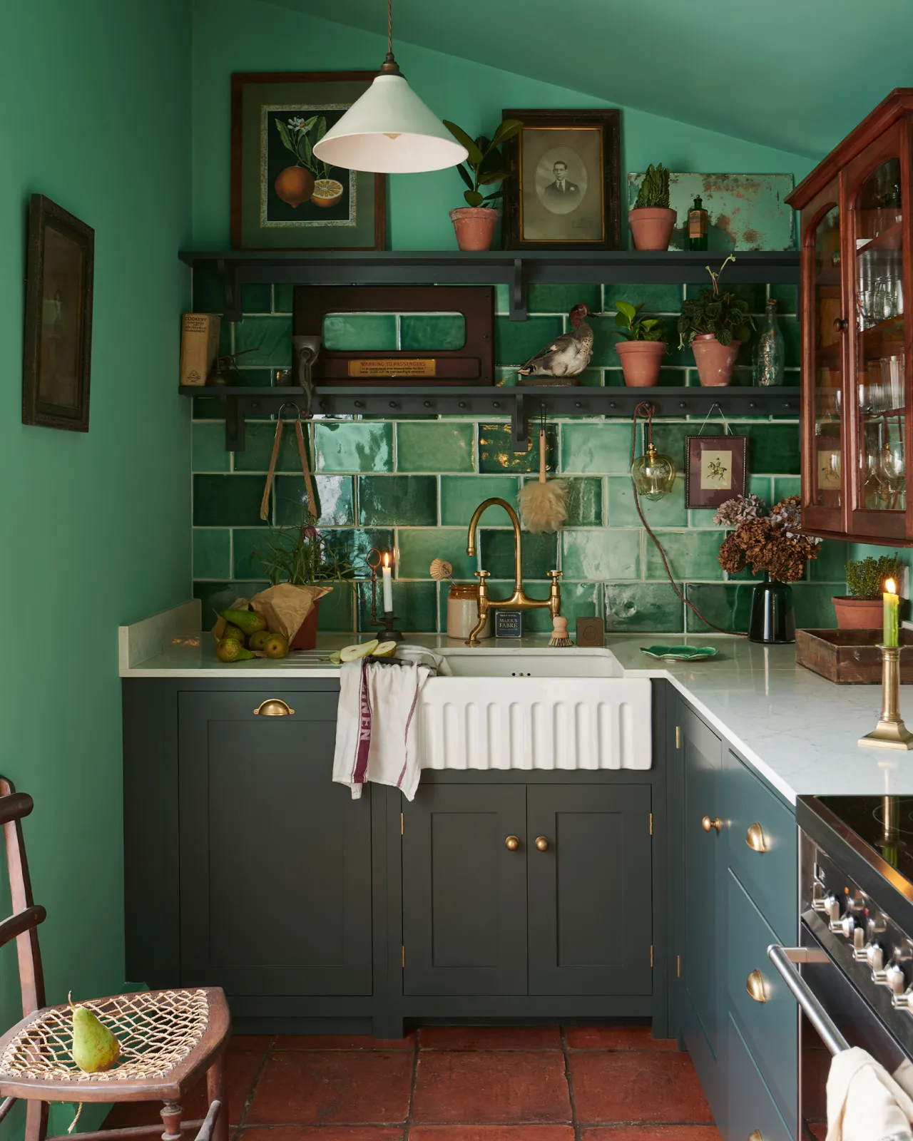 An eclectic cottage kitchen with dark green painted Shaker-style cupboards, arsenic green walls, glossy emerald green wall tiles, vintage bric-a-brac and a terracotta tiled floor.