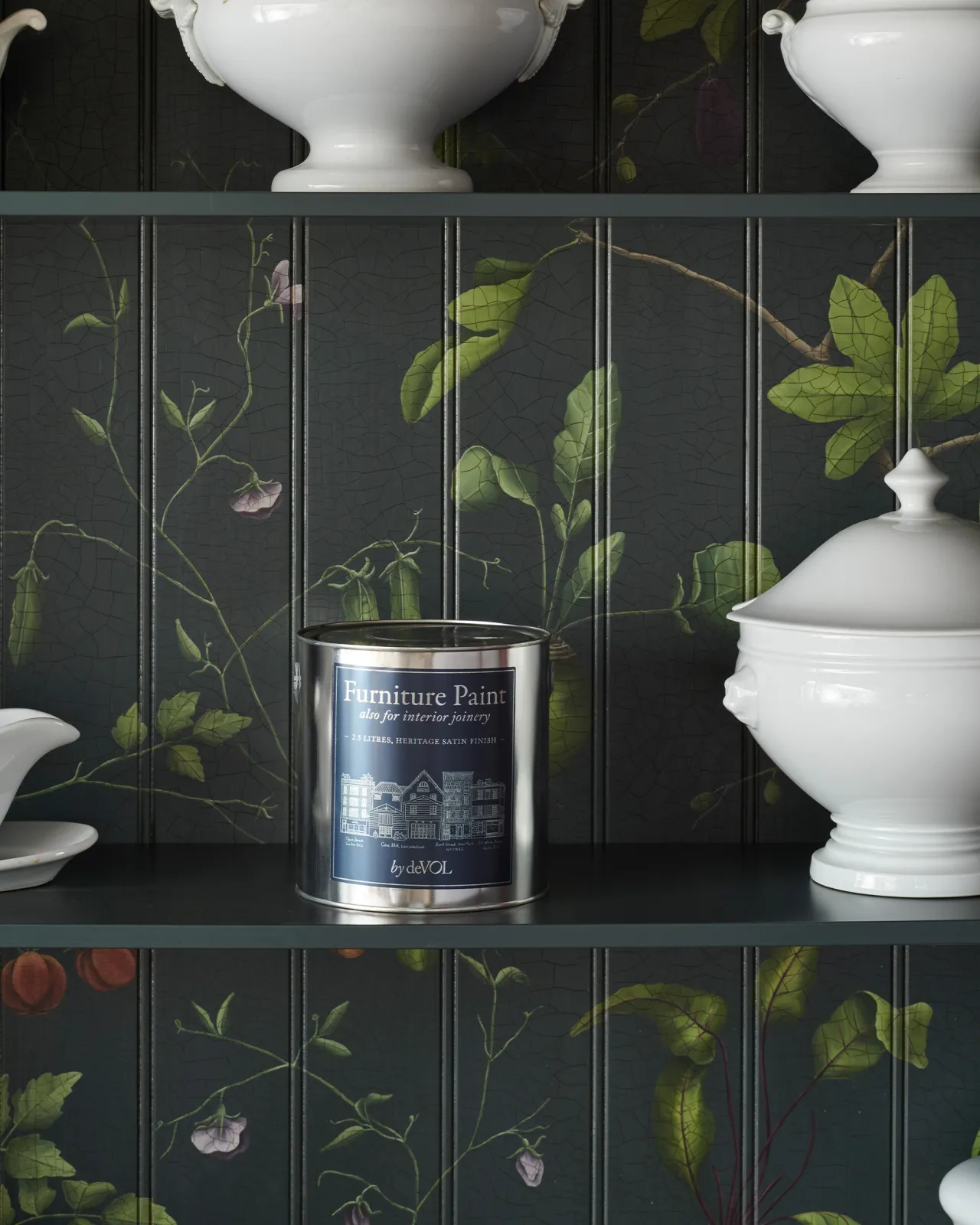 A silver paint tin with a rich blue label inside a dark green cupboard interior with hand-painted botanical flora on a tongue and groove-style backboard.
