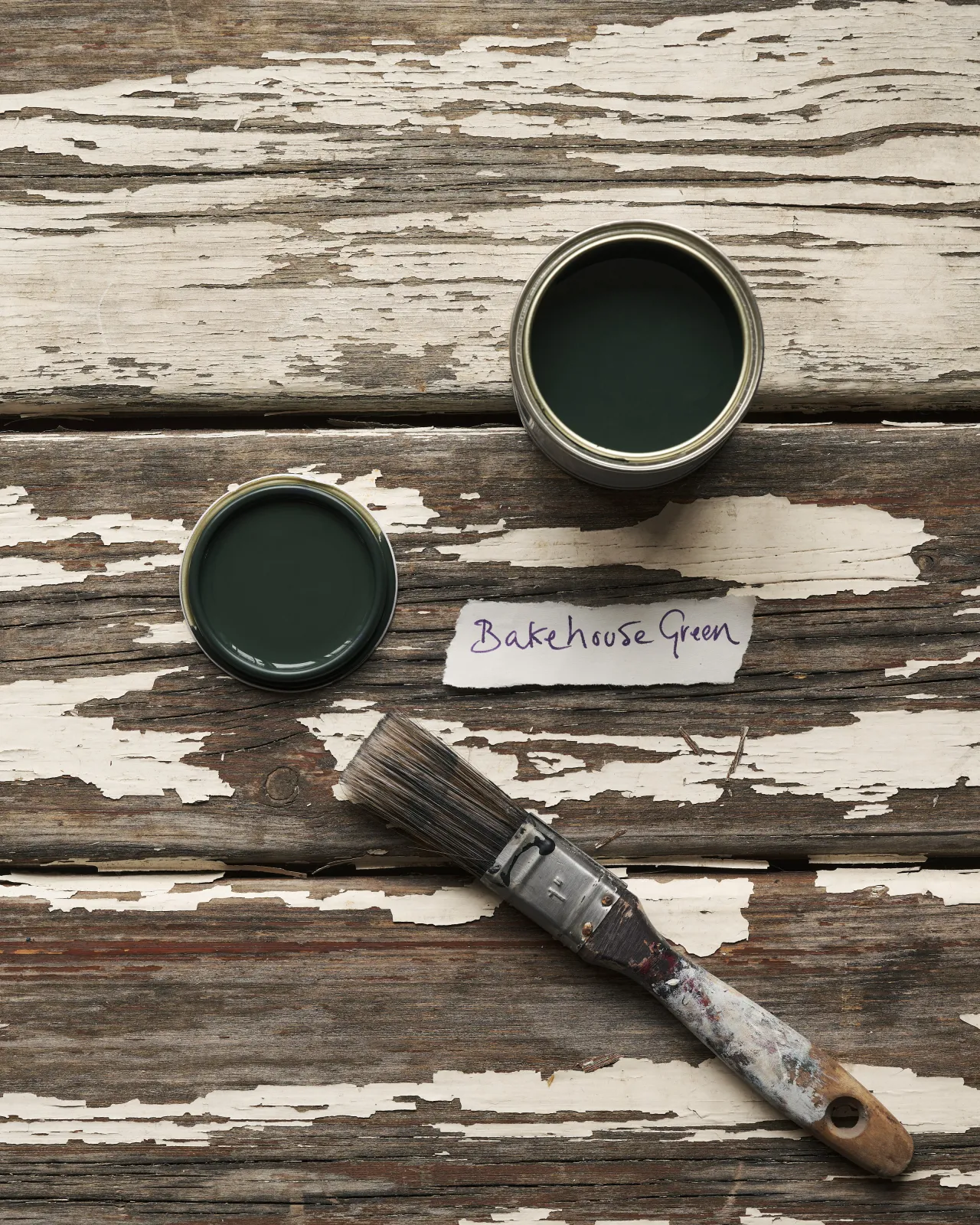 An overhead view of an open paint pot and a worn brush on a rustic tabletop. The dark green colour inside is labelled 'Bakehouse Green'.