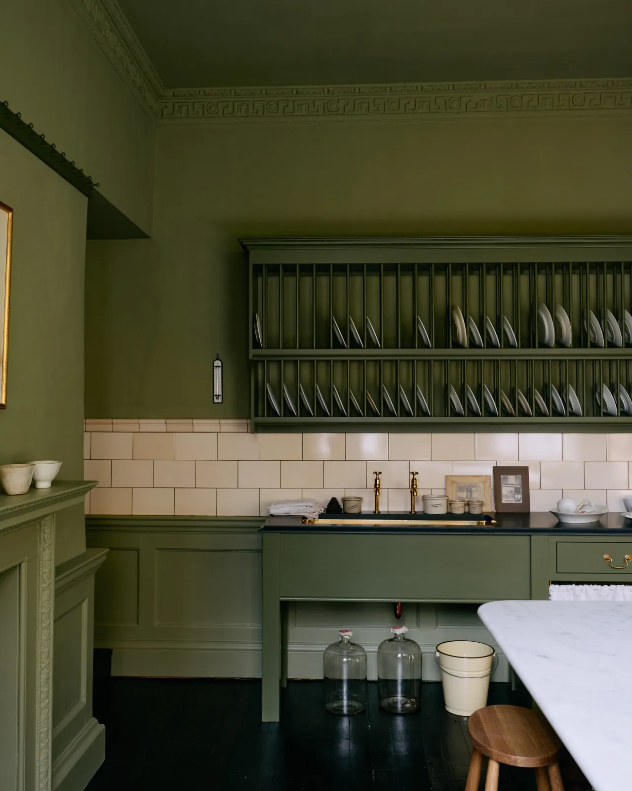 A splashback band of simple cream subway wall tiles stretches across a moody green room with a traditional Victorian-style kitchen.