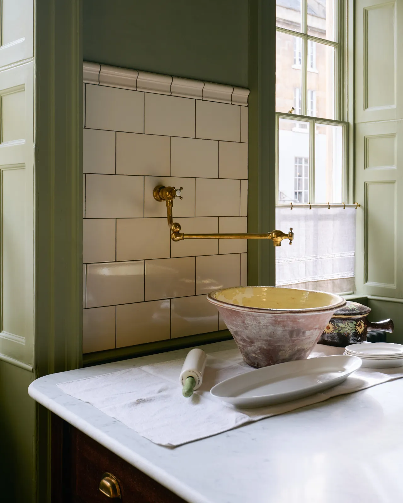 A section of wall between two tall original Georgian sash windows is tiled with simple glossy cream subway tiles. An aged brass pot filler tap extends from the wall ready to fill a large ceramic bowl on the table below.