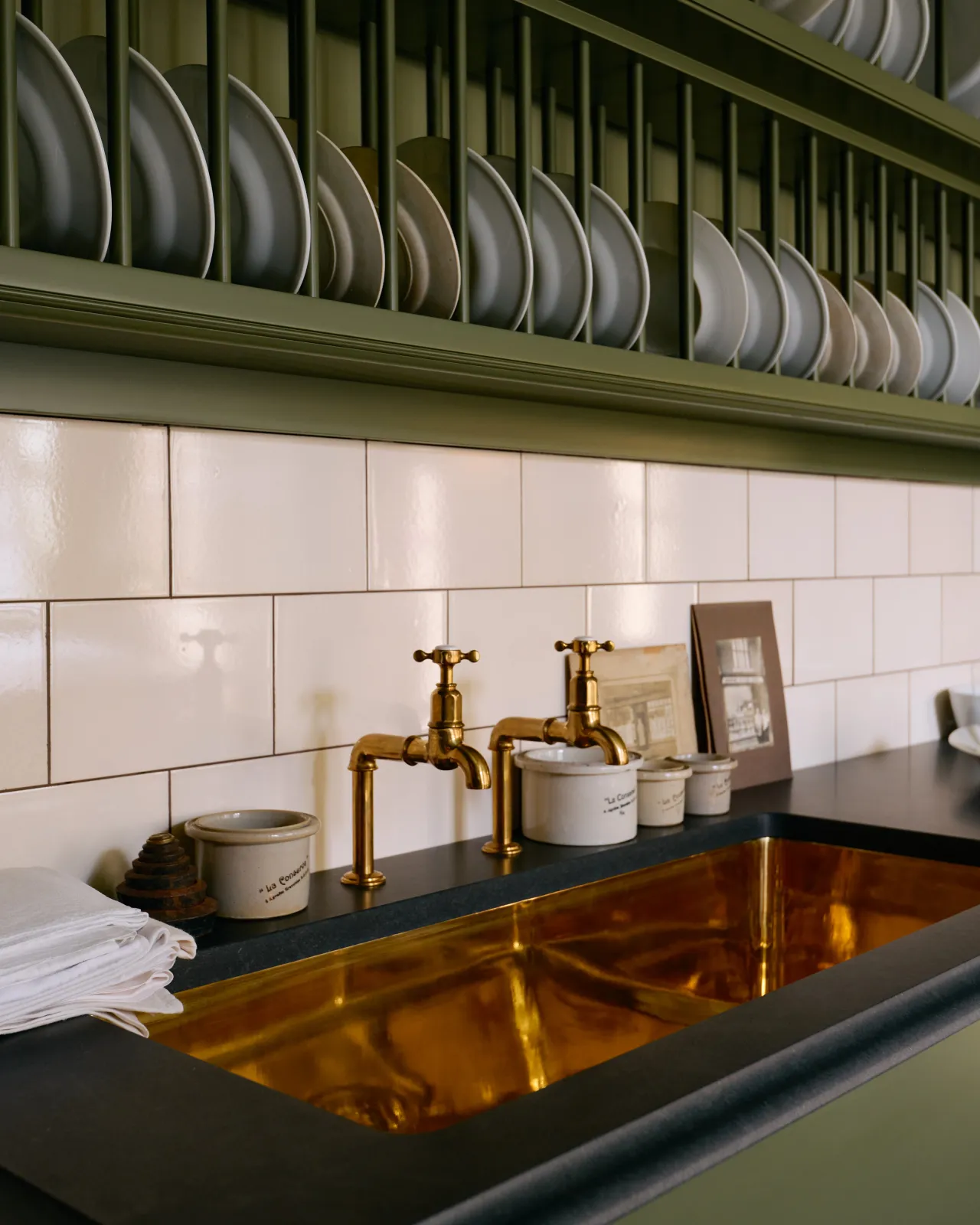 An oblique view of a grand polished brass sink with aged brass bibcock taps. A splashback of simple cream subway wall tiles runs behind the taps.
