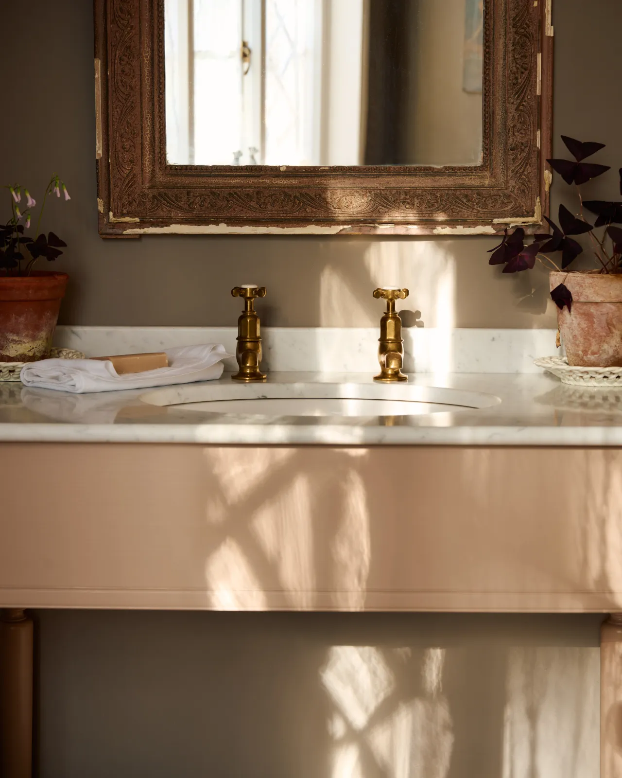 A straight-on view of a pair of aged brass pillar taps with crosshead handles fitted to a pink painted Victorian-inspired bathroom washstand.