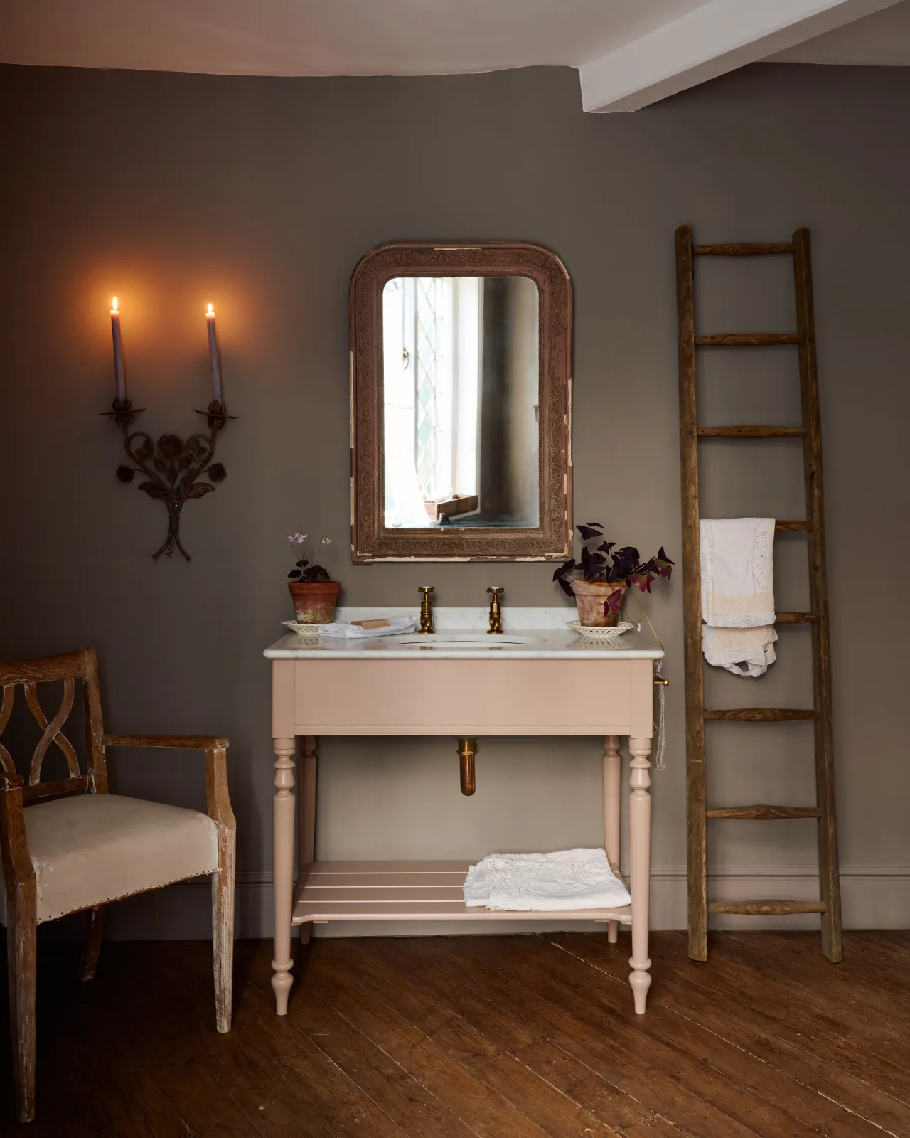 A straight-on view of a dusty-pink Victorian-inspired washstand with brass pillar taps in a greyish-brown painted room. The shot is atmospheric and moody, with two lit candles casting a subtle glow and light from an adjacent window illuminating the brass accents.