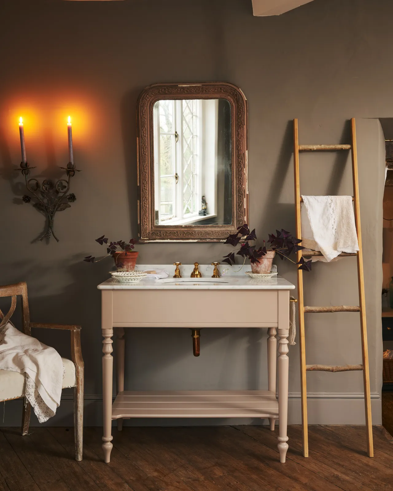A straight-on view of a dusty-pink Victorian-inspired washstand with a brass mixer tap in a greyish-brown painted room. The shot is atmospheric and moody, with two lit candles casting a subtle glow and light from an adjacent window illuminating the brass accents.