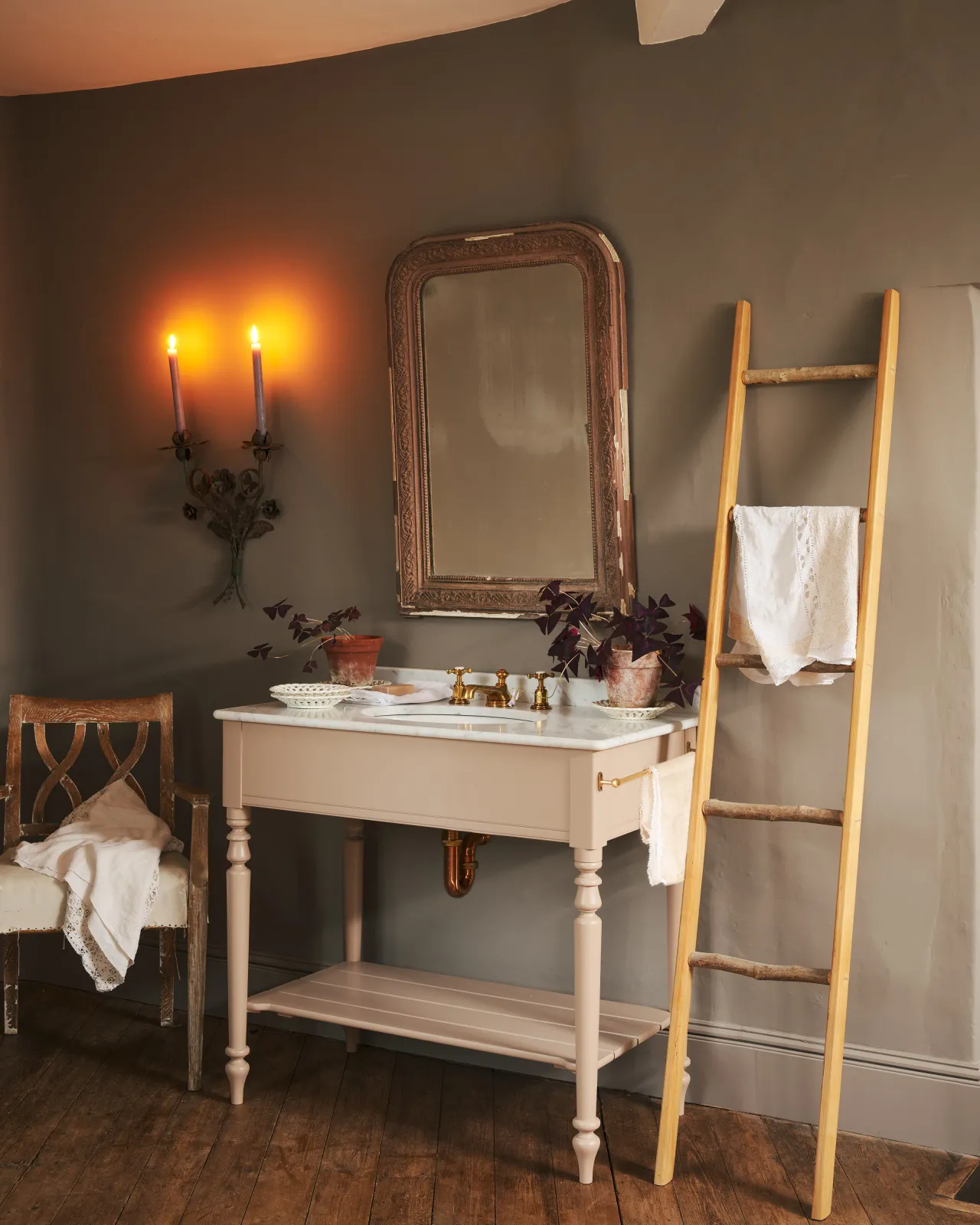 An oblique view towards a dusty-pink Victorian-inspired washstand with a brass mixer tap in a greyish-brown painted room. The shot is atmospheric and moody, with two lit candles casting a subtle glow and light from an adjacent window illuminating the brass accents.