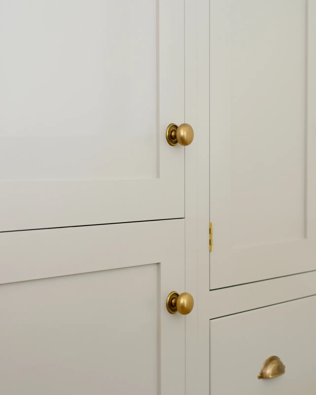 An oblique view of a cream painted Shaker-style pantry cupboard with a set of round doorknobs in a burnished 'Bella' brass finish.