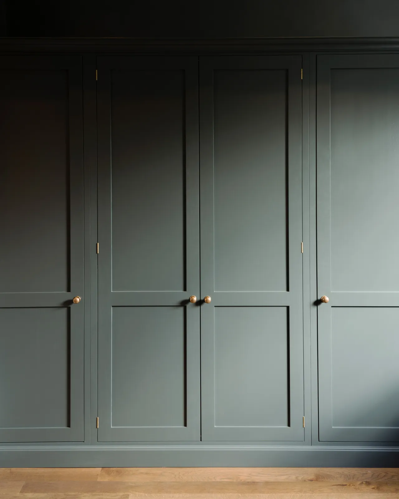 A straight-on view of dark sage green painted Shaker-style full height pantry cupboards, fitted with round doorknobs in a burnished 'Bella' brass finish.