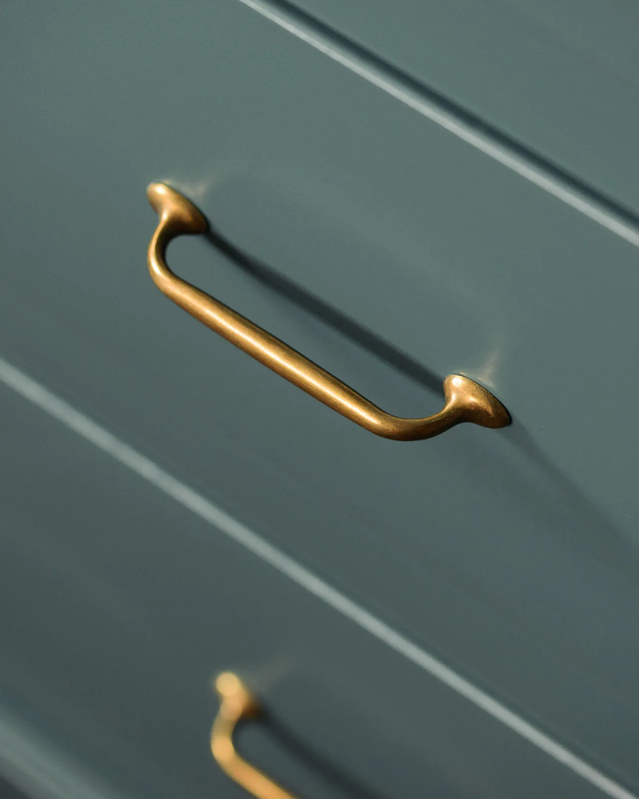 Simple pull handles in a burnished 'Bella' brass finish fitted to blue-green painted cupboard drawers.