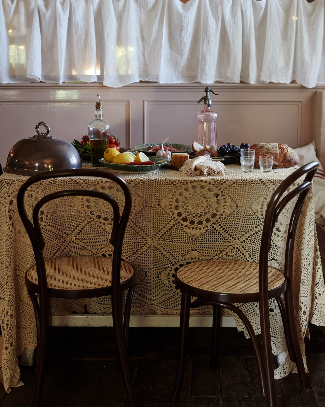 Two café-style bentwood chairs with dark stained frames and light woven rattan seat mesh in front of a table laid with a vintage lace tablecloth and platters of food.