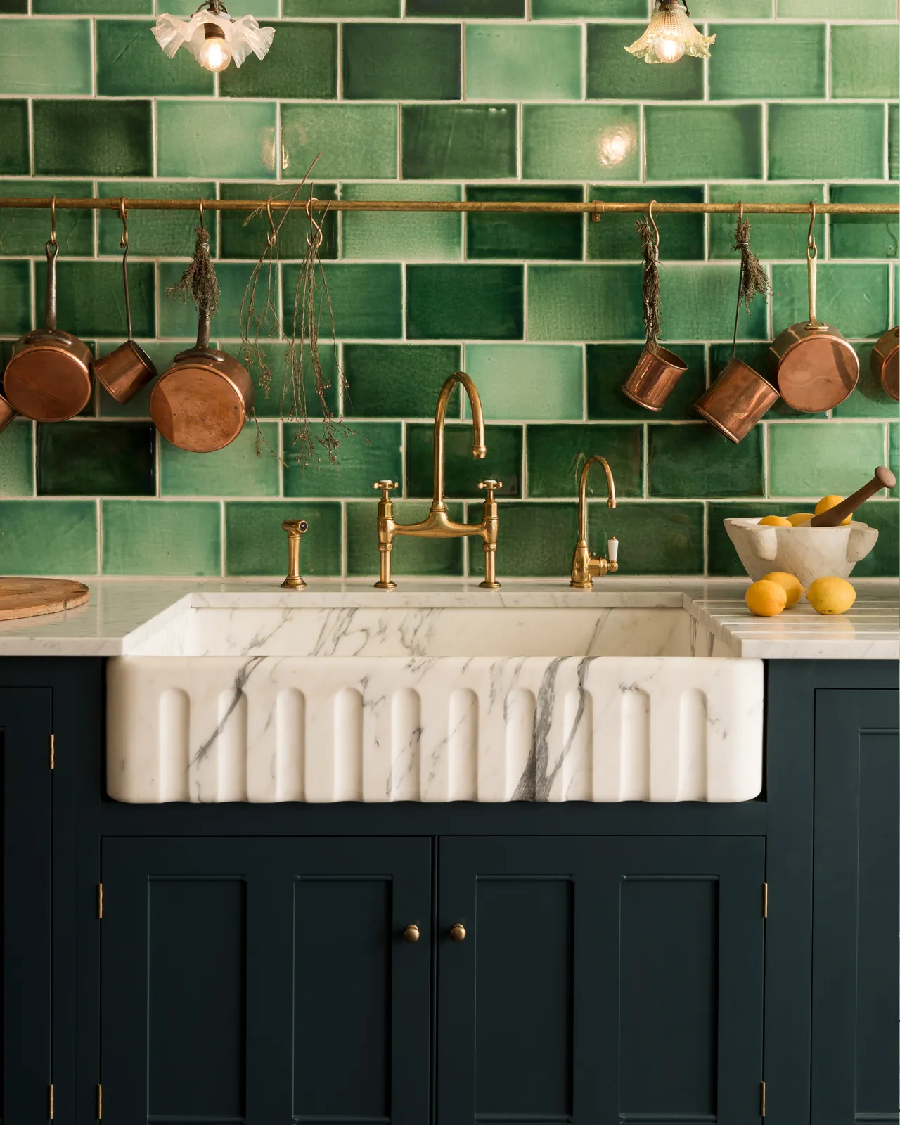 A straight-on view of a grand Arabescato marble sink with a fluted front, with royal blue-coloured Georgian-style kitchen cupboards, aged brass taps and a wall of glossy emerald green tiles.