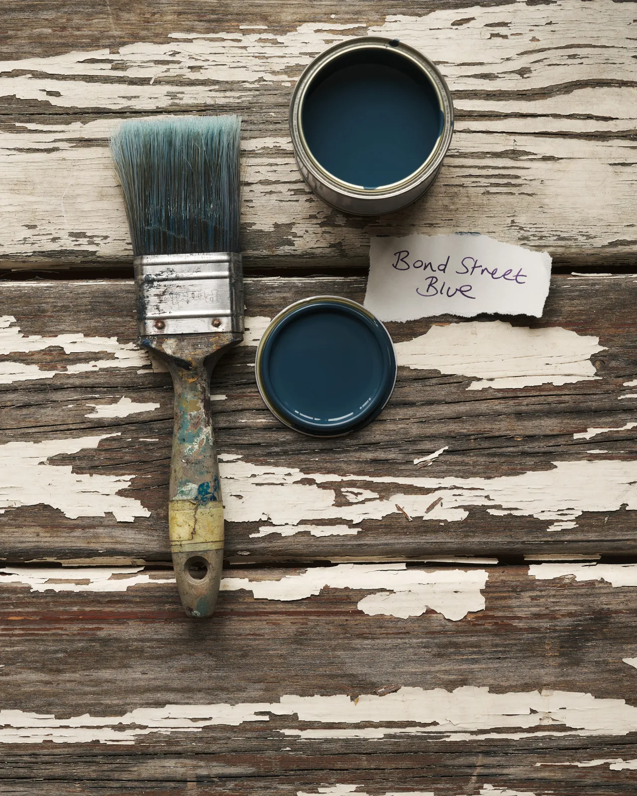 An overhead view of an open paint pot and a worn brush on a rustic tabletop. The royal blue colour inside is labelled 'Bond Street Blue'.