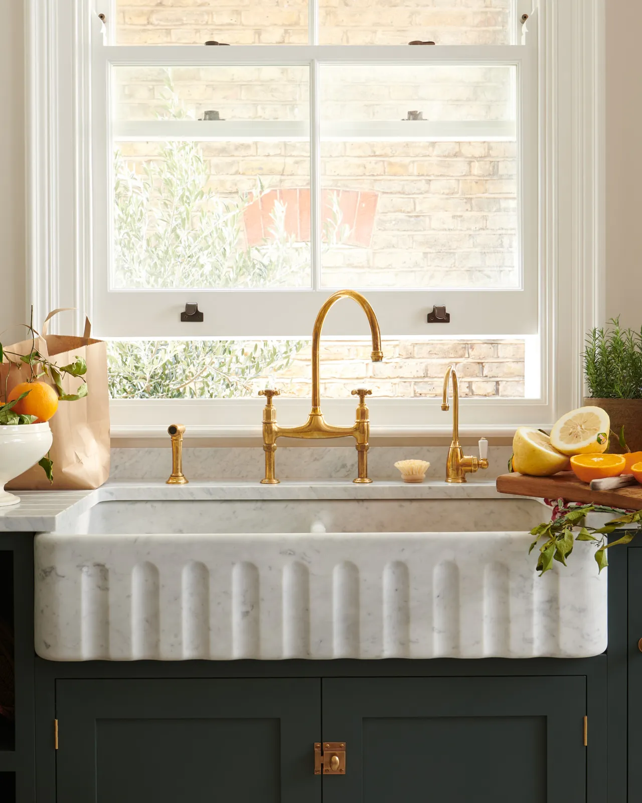 A straight-on view of a small traditional-style aged brass tap with a white ceramic lever, seen with a fluted Carrara marble sink and a larger aged brass mixer tap and an independant rinser tap.