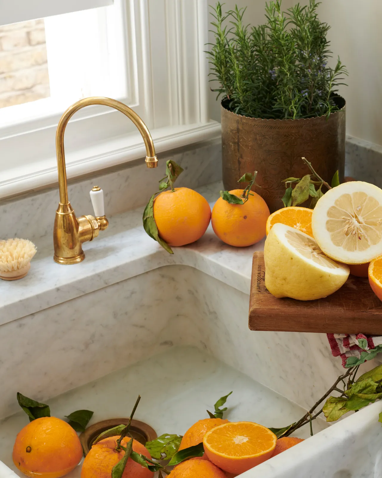 A small traditional-style aged brass tap with a white ceramic lever above a Carrara marble sink and a chopping board littered with orange slices.