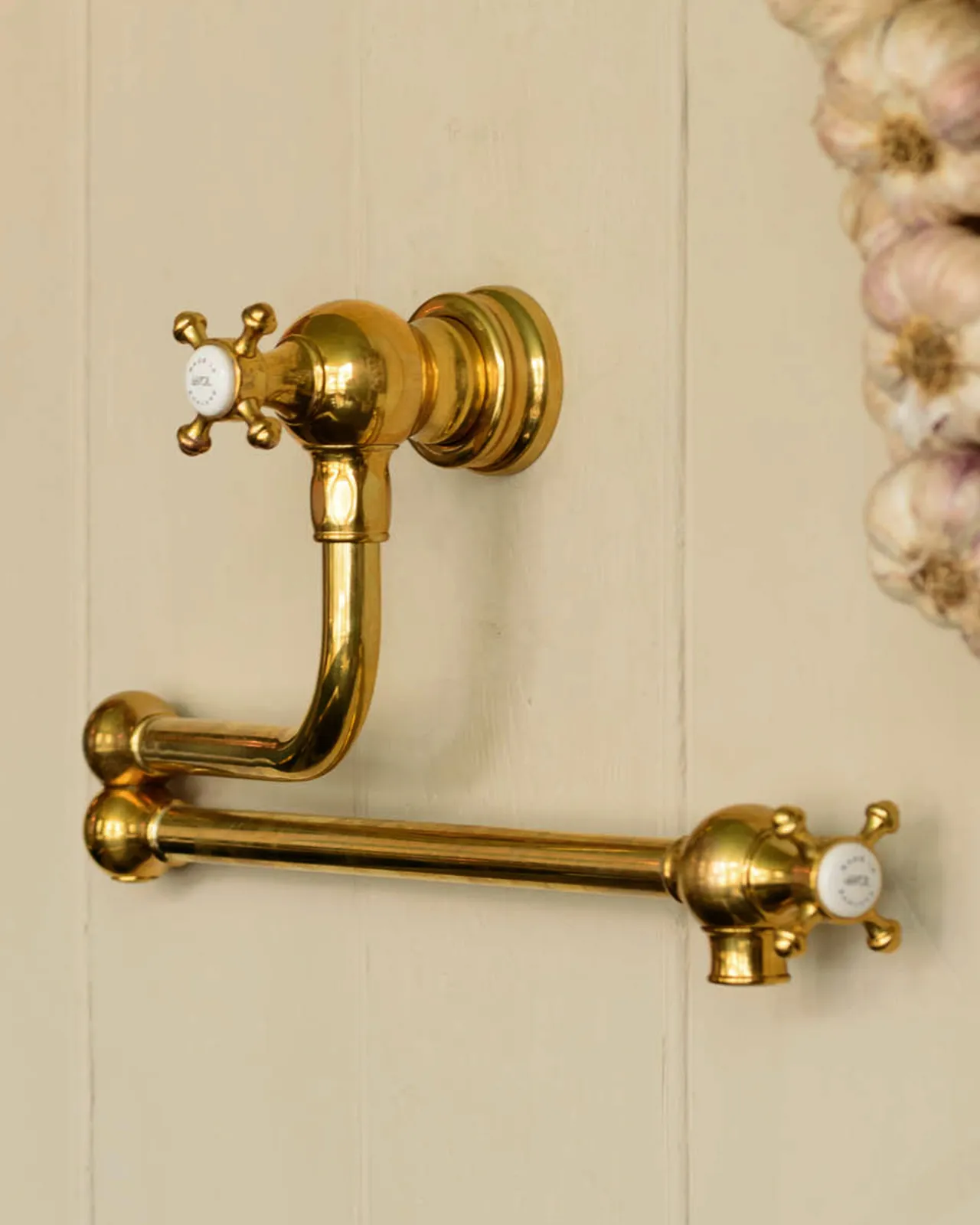 An oblique view of a traditional-style aged brass pot filler tap with crosshead handles fitted to a creamy coloured tongue and groove panelled wall.