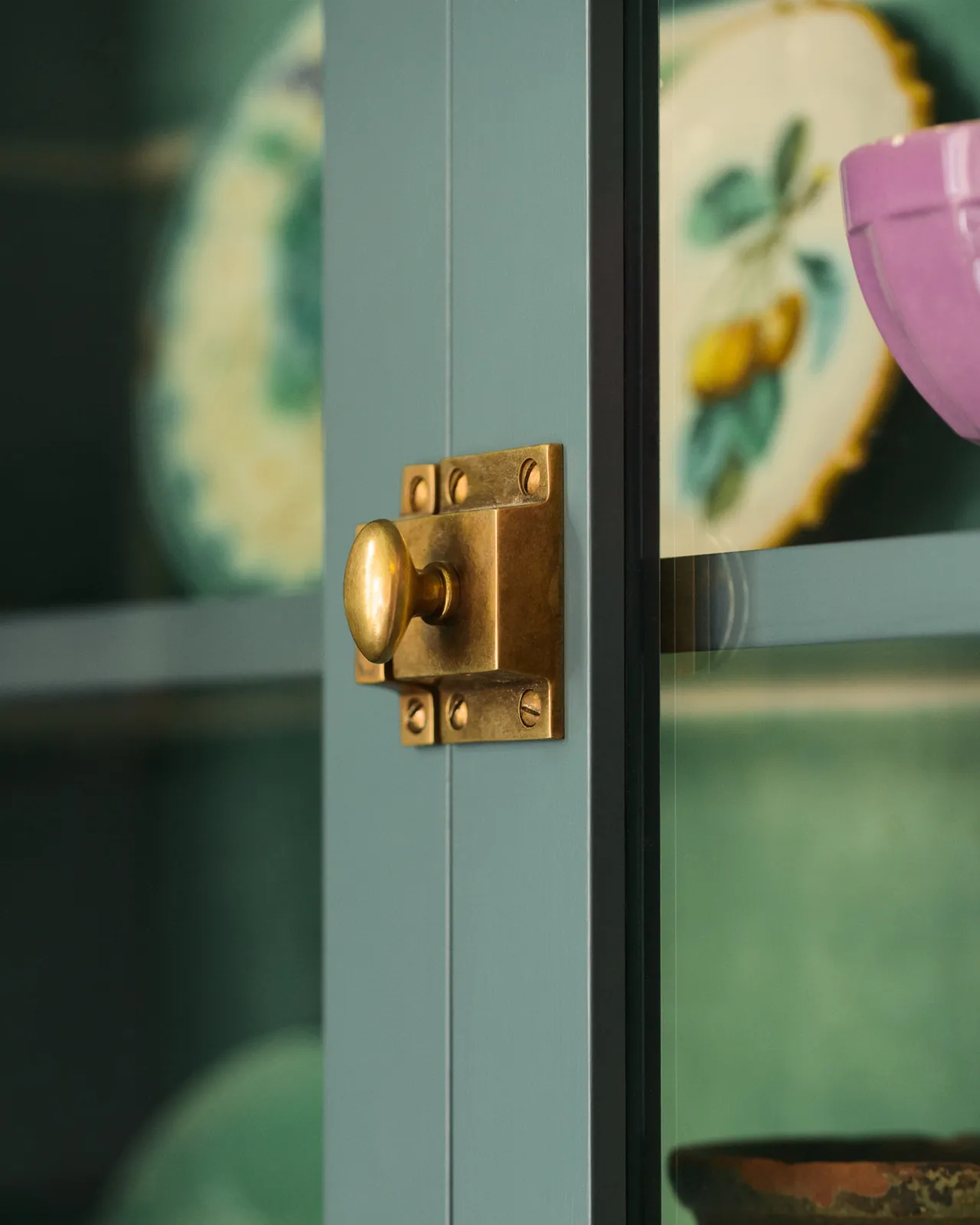 An oblique view of a square cupboard catch with an oval-shaped knob in a burnished 'Bella' brass finish fitted to a glazed kitchen cupboard door with a blue-green painted frame.