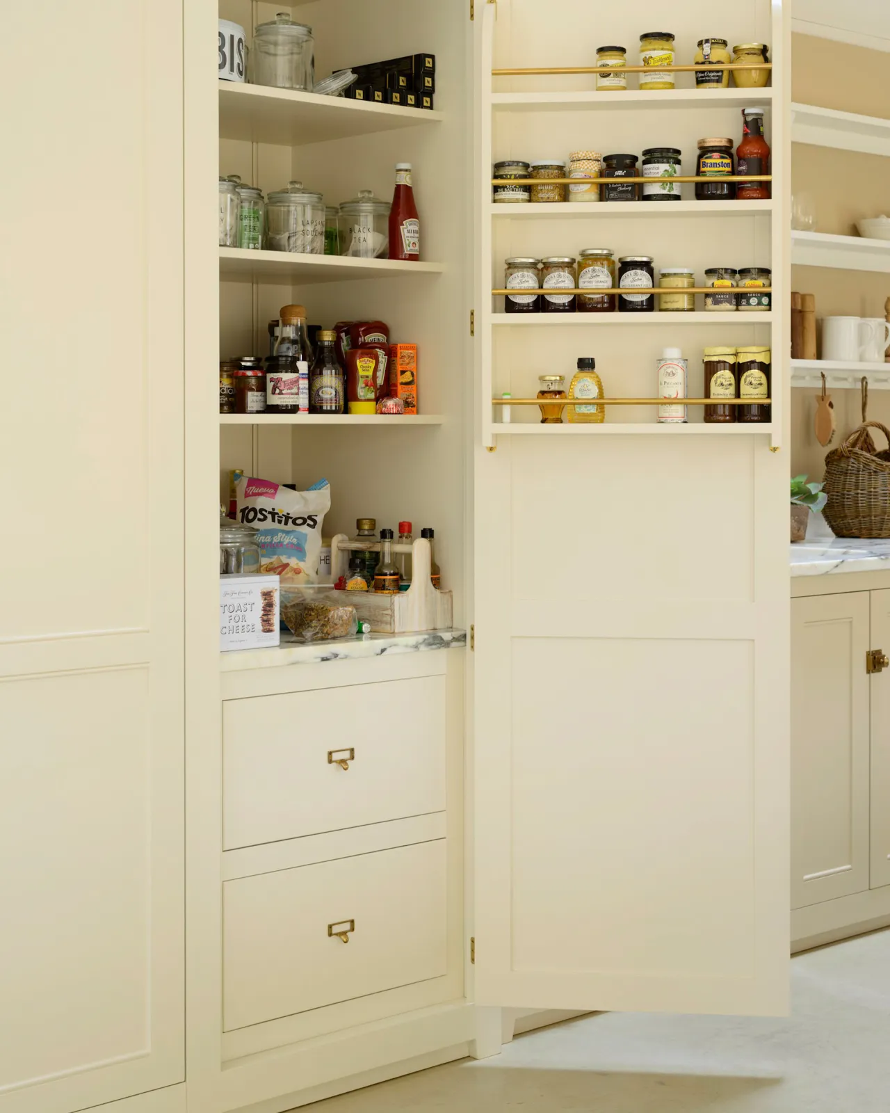 An oblique view of a tall fitted pantry painted in a cream colour. One door is open to reveal two internal drawers with small cardframe pull handles in a burnished 'Bella' brass finish.