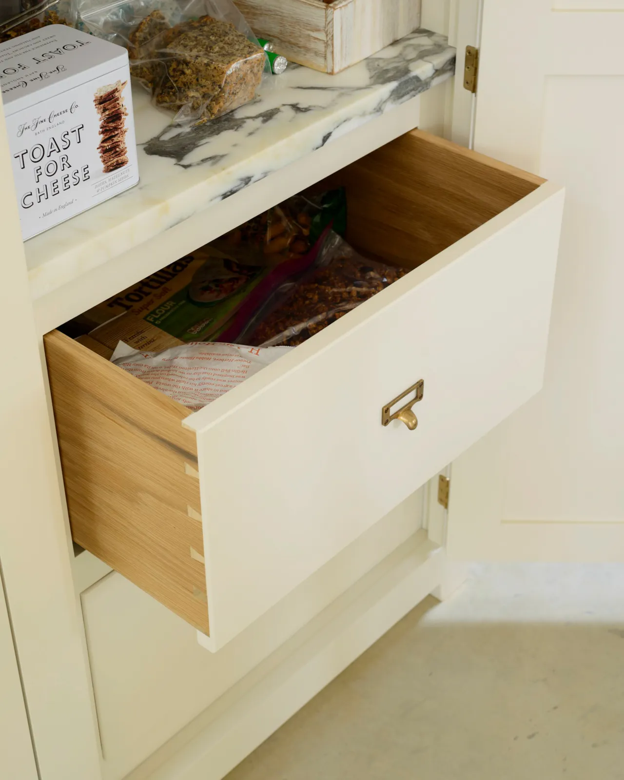 An oblique view of a small cardframe pull handle in a burnished 'Bella' brass finish fitted to an oak drawer box with a cream painted drawer front.