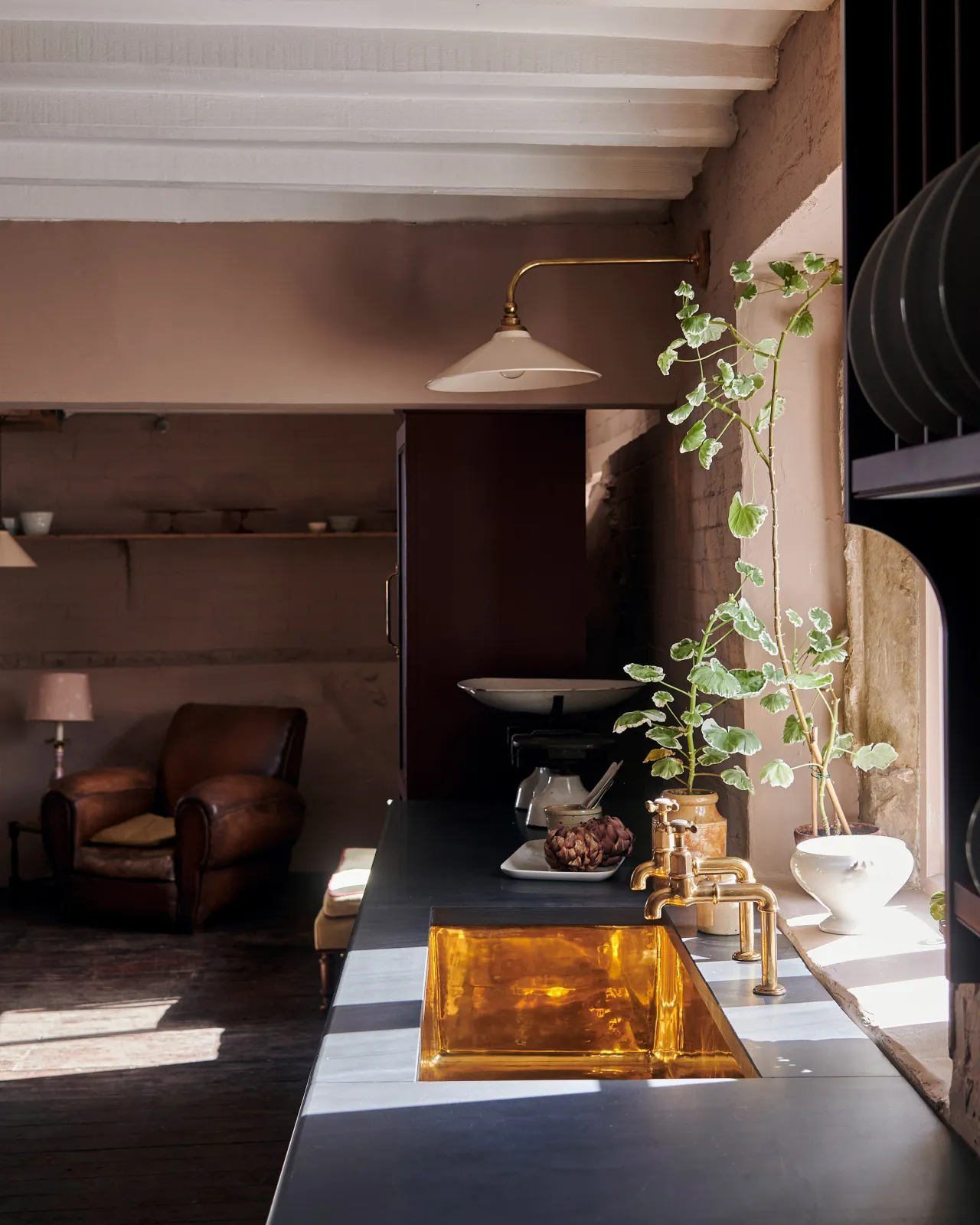 Looking across a slate kitchen worktop with a large undermounted polished brass sink with aged brass bibcock taps and a tall geranium plant.