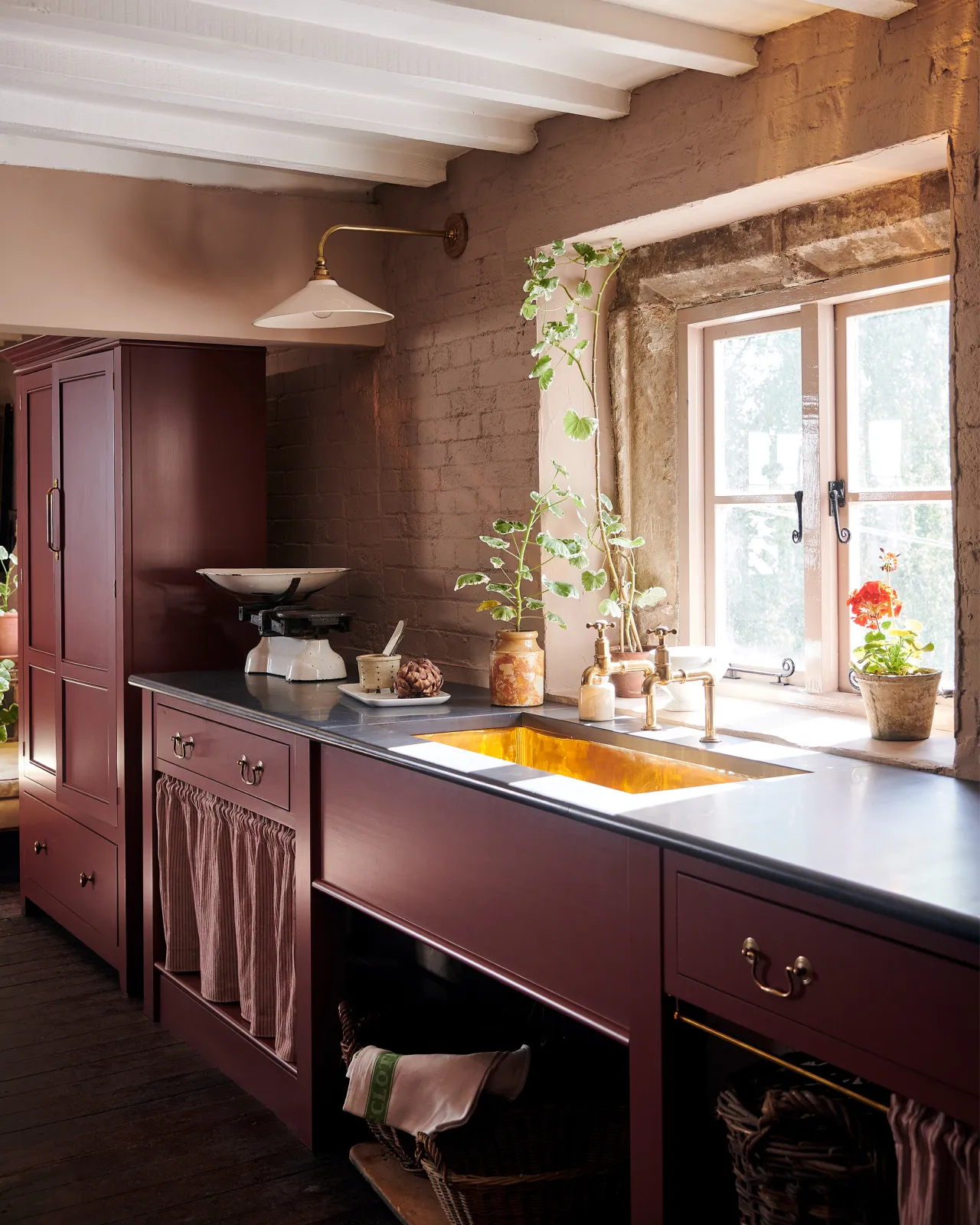 An oblique view of a red painted Victorian-style kitchen sink cupboard with a slate worktop and a large undermounted polished brass sink.