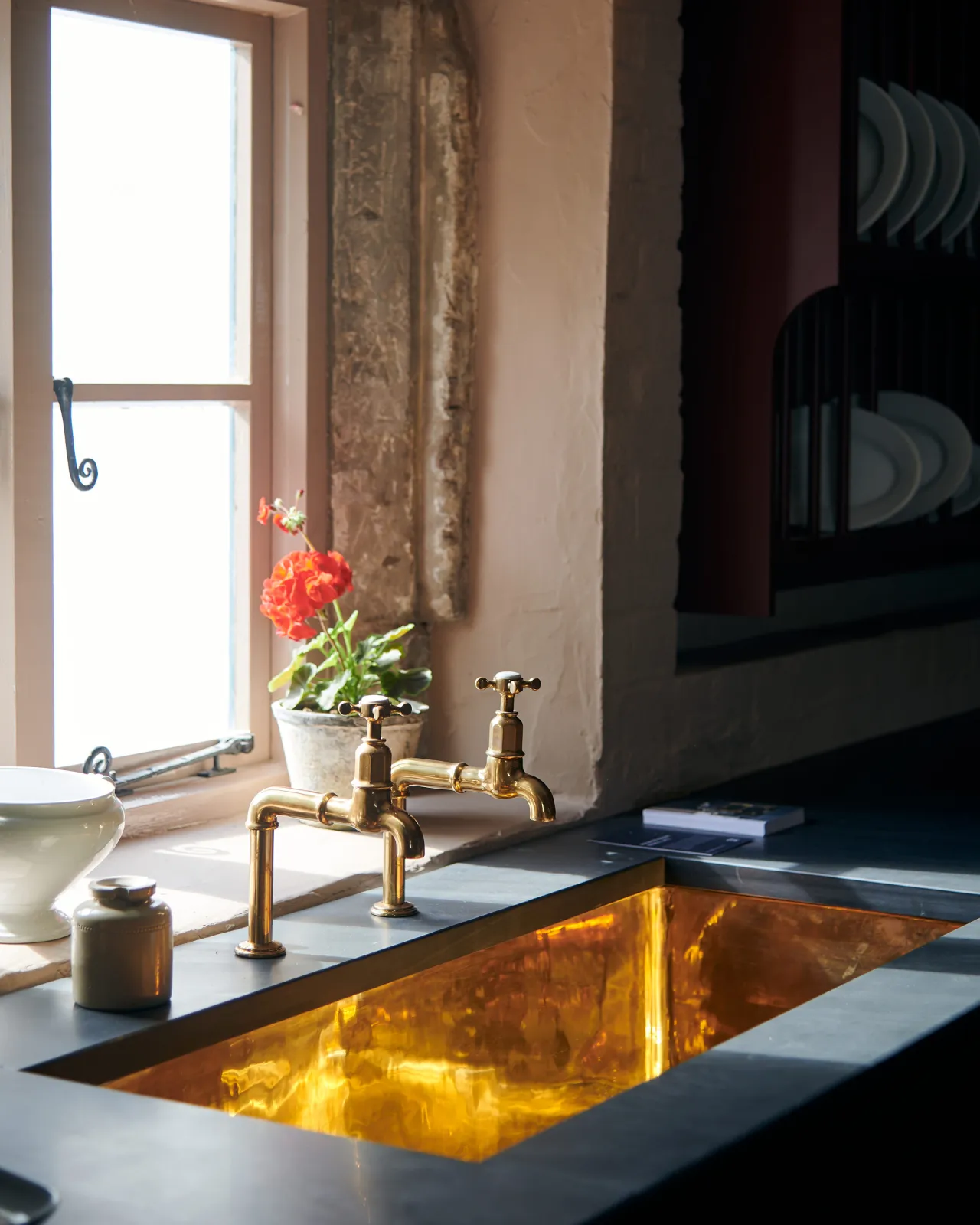 An oblique view of a large undermounted polished brass sink with aged brass bibcock taps and a tall geranium plant.