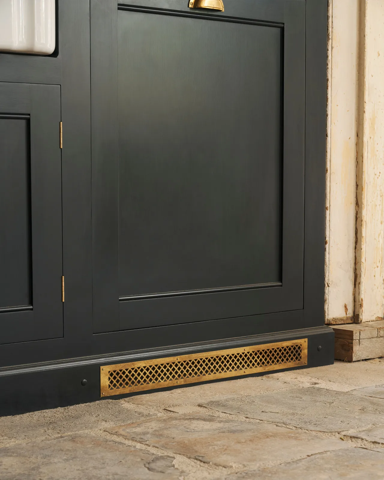 An oblique view of a black painted Georgian-style kitchen cupboard with a classic brass ventilation cover fitted to the skirting.