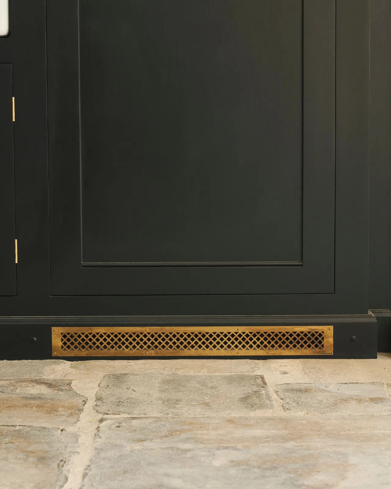 A straight-on view of a black painted Georgian-style kitchen cupboard with a classic brass ventilation cover fitted to the skirting.