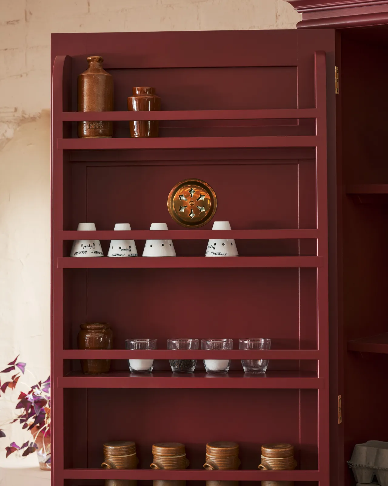 The inside of a red painted pantry cupboard door with a spice rack and the decorative brass backplate of a Victorian-inspired circular ventilation grille.