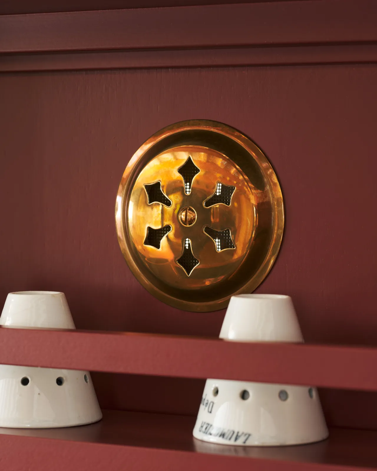 A close-up oblique view of a Victorian-inspired circular brass ventilation grille with traditional decorative cutouts fitted to a red painted cupboard door.