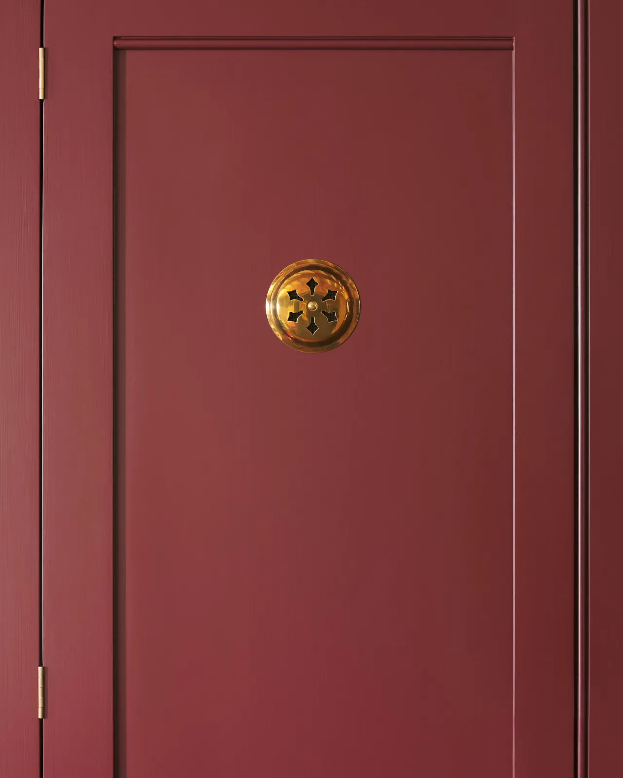A Victorian-inspired circular brass ventilation grille with traditional decorative cutouts fitted to a red painted cupboard door.