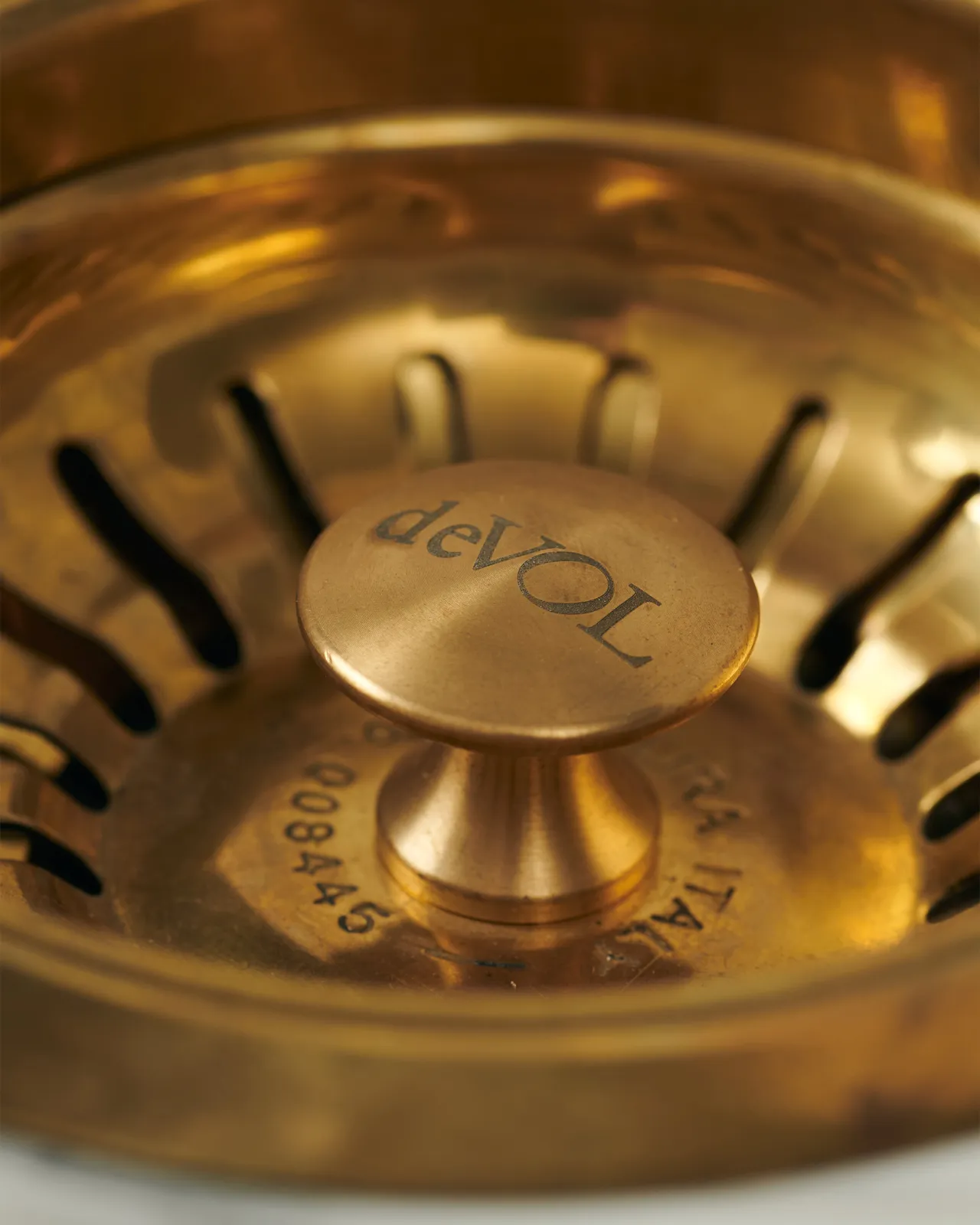 A close-up view of the central finial of a warm burnished brass basket waste strainer, there is a small 'deVOL' logo in the middle of the finial.
