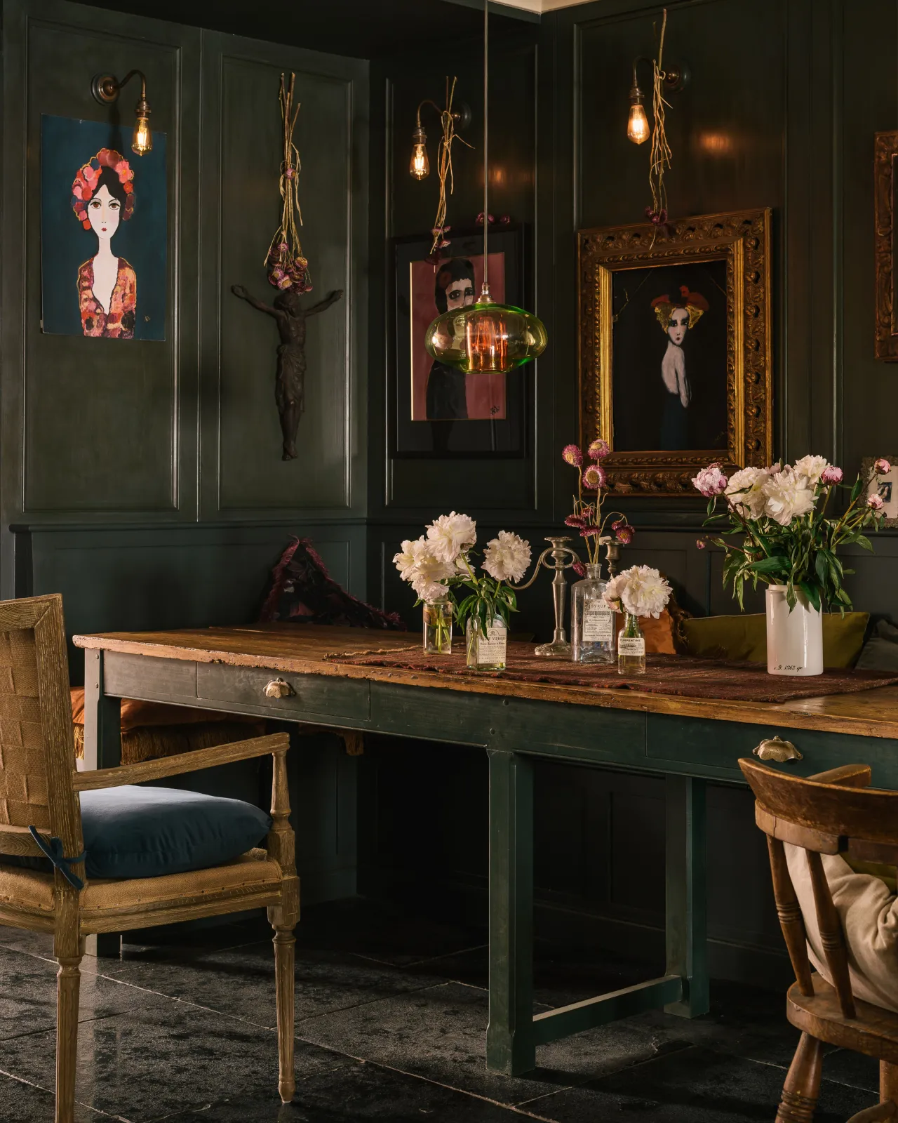 An oblique look towards an atmospheric dining area, with a long rustic table, antique chairs and built-in bench seating. Dark green walls and furniture, and Brushed Charcoal Limestone floors create a moody, aged feel.