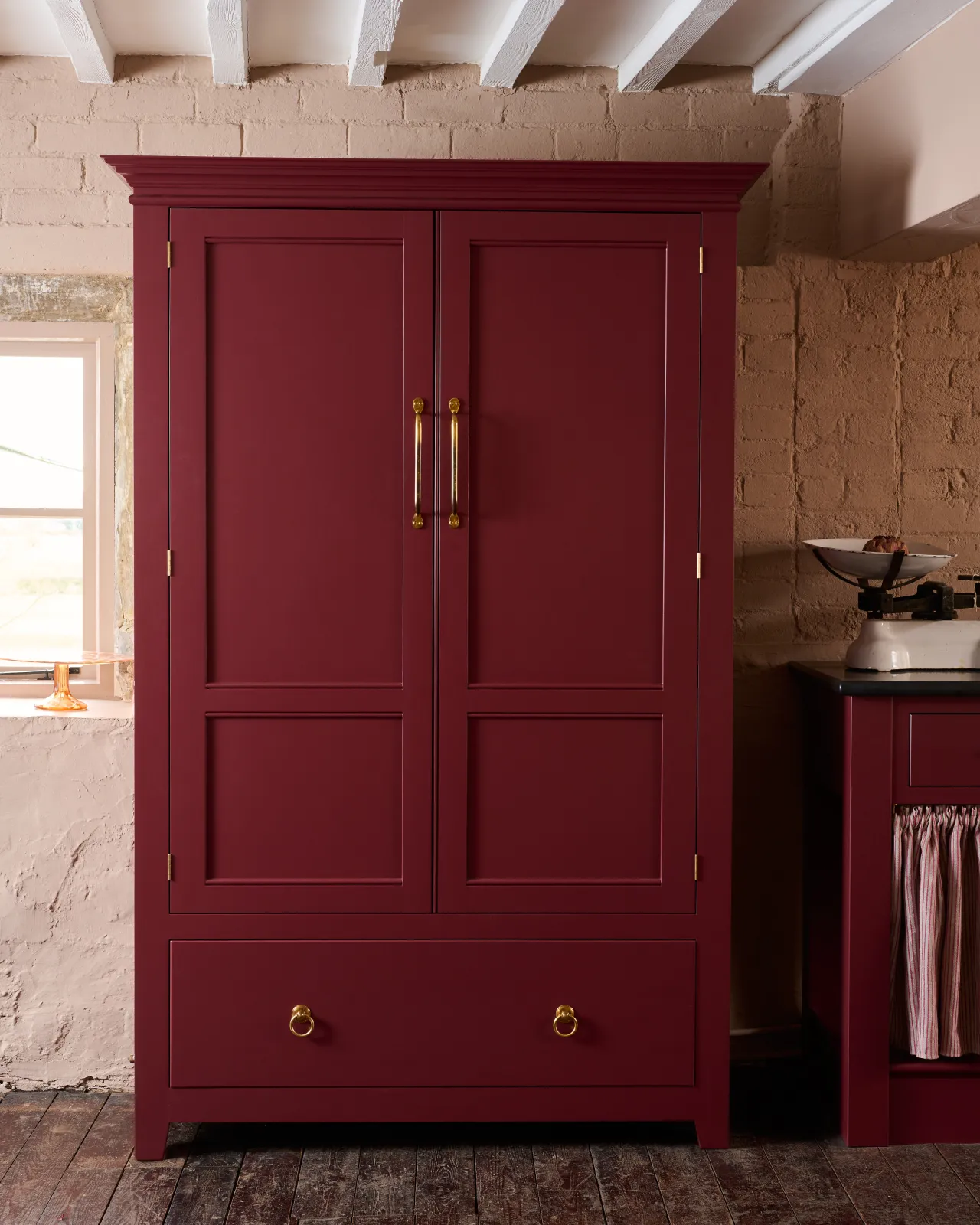 A tall red painted Georgian-style pantry cupboard with double doors and a bottom drawer, fitted with Victorian-inspired ring pull handles in a warm burnished brass finish.