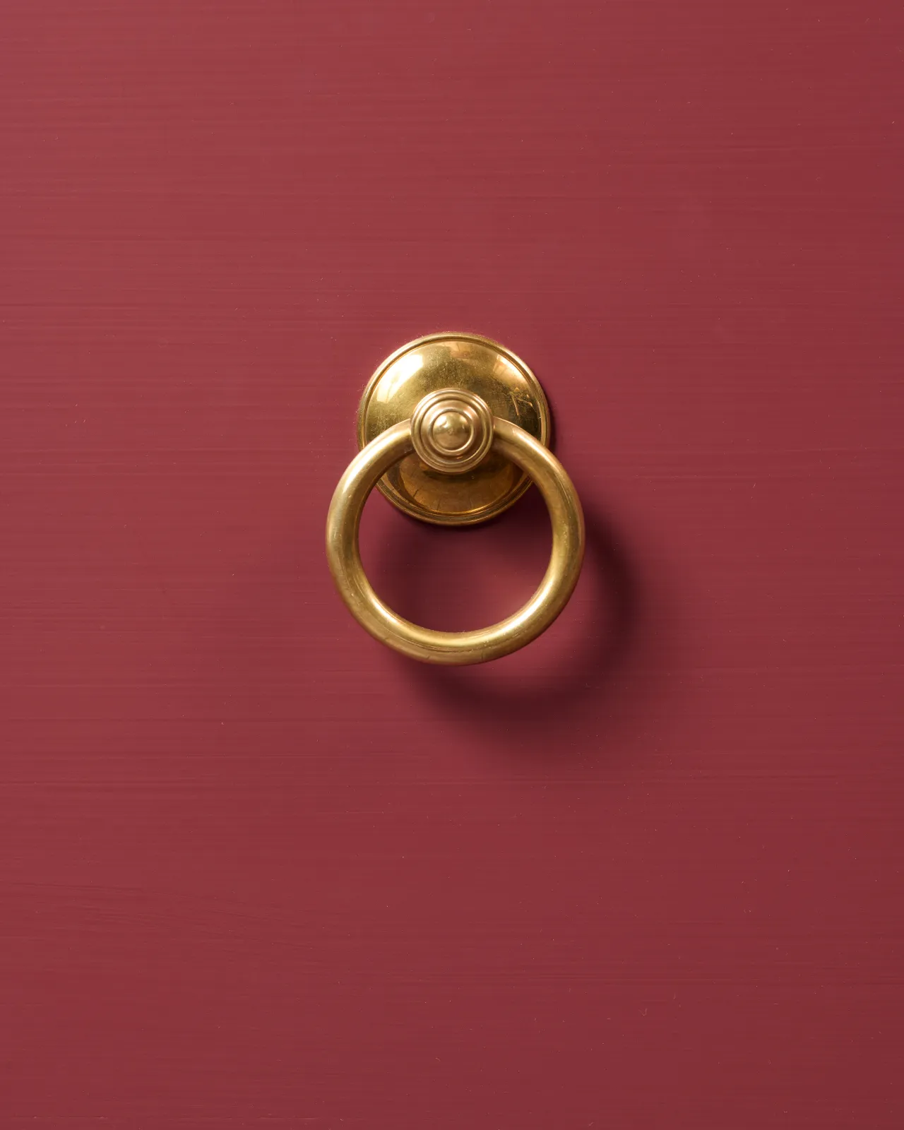 A close-up straight-on view of a Victorian-inspired ring pull handle in a warm burnished brass finish, fitted to a red painted cupboard drawer.