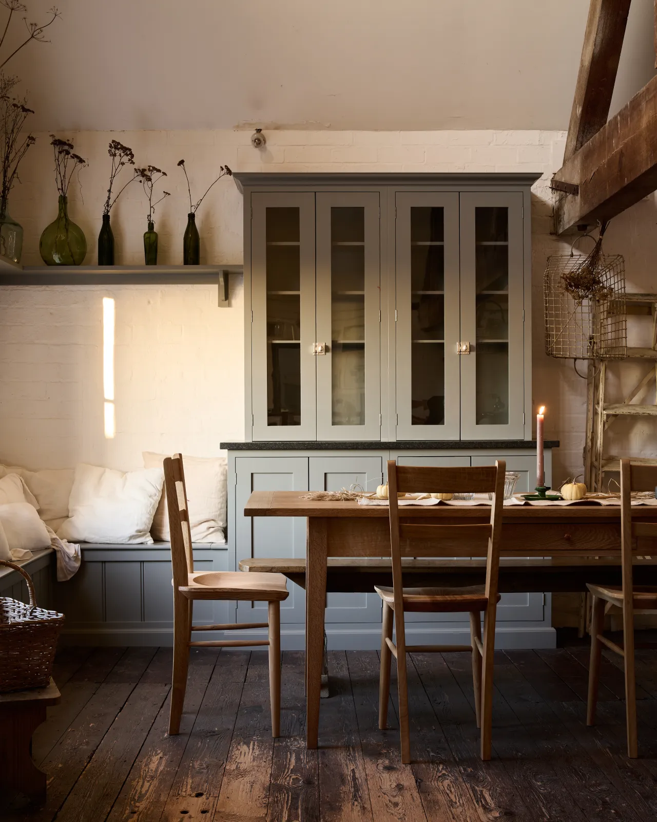 A straight-on view of a rustic kitchen with worn floorboards, white brick walls and grey painted cabinetry. A large table is surrounded by mid-century-style dining chairs with sculpted seat pans.