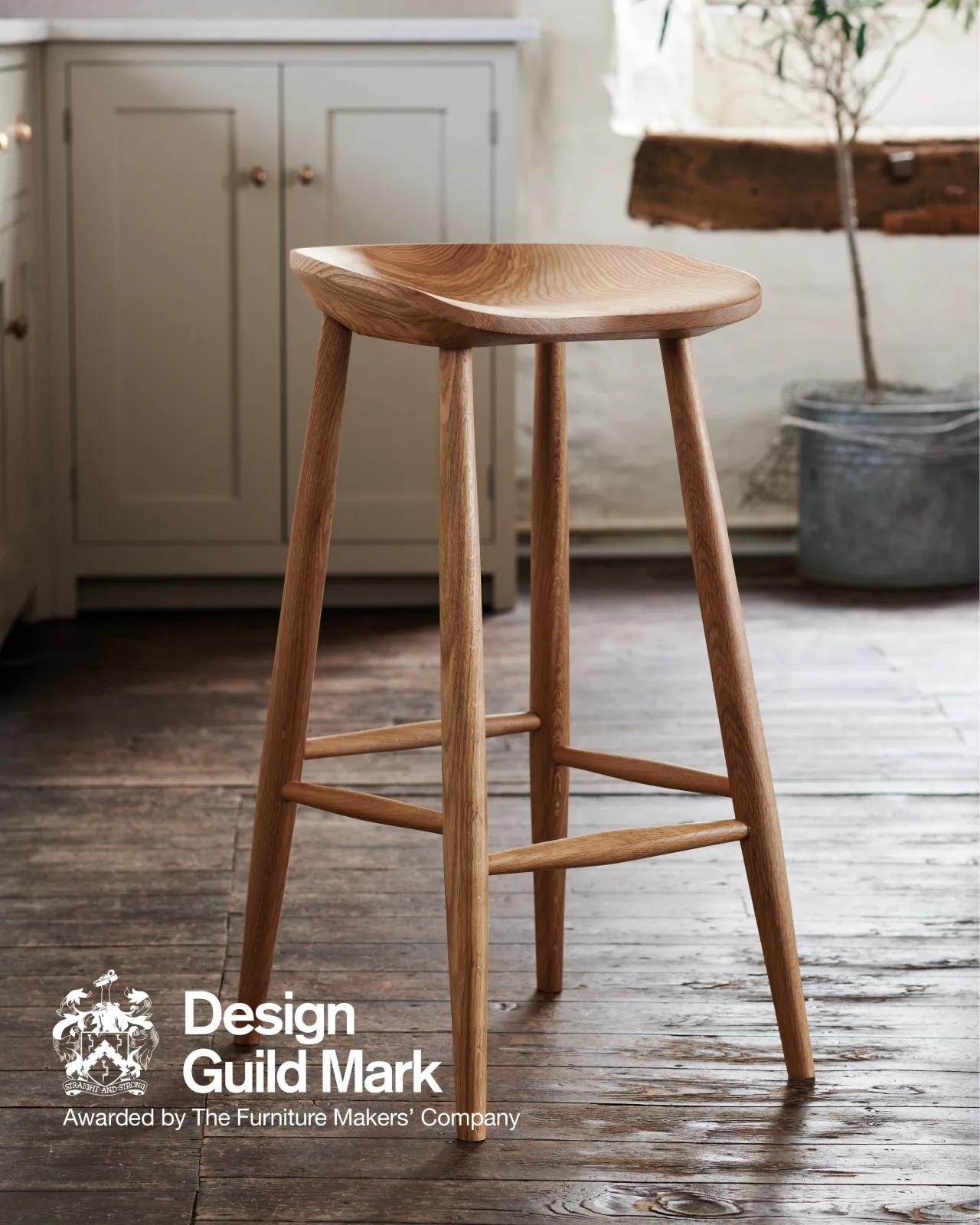 A mid-century-style bar stool with a sculpted ash seat pan and oak legs. A logo in the bottom corner reads 'Design Guild Mark; Awarded by the Furniture Makers' Company'.