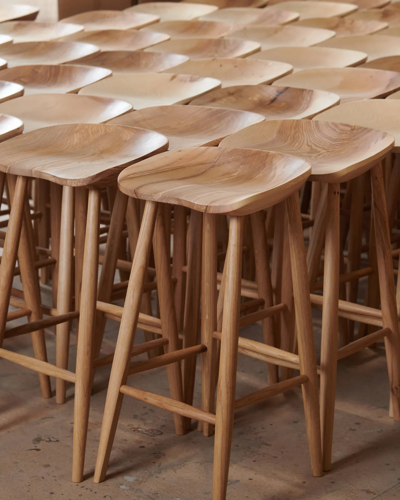An oblique view of a large set of mid-century-style bar stools with a sculpted ash seat pans and oak legs. The wood grain of each seat pan is slightly different in tone and pattern.