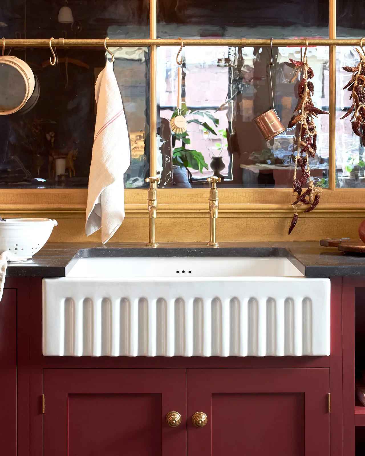 A close-up straight-on view of a Shaker-style kitchen sink cupboard with a fluted Belfast sink, red painted cupboard doors and Victorian-inspired ornate doorknobs in a warm burnished brass finish.