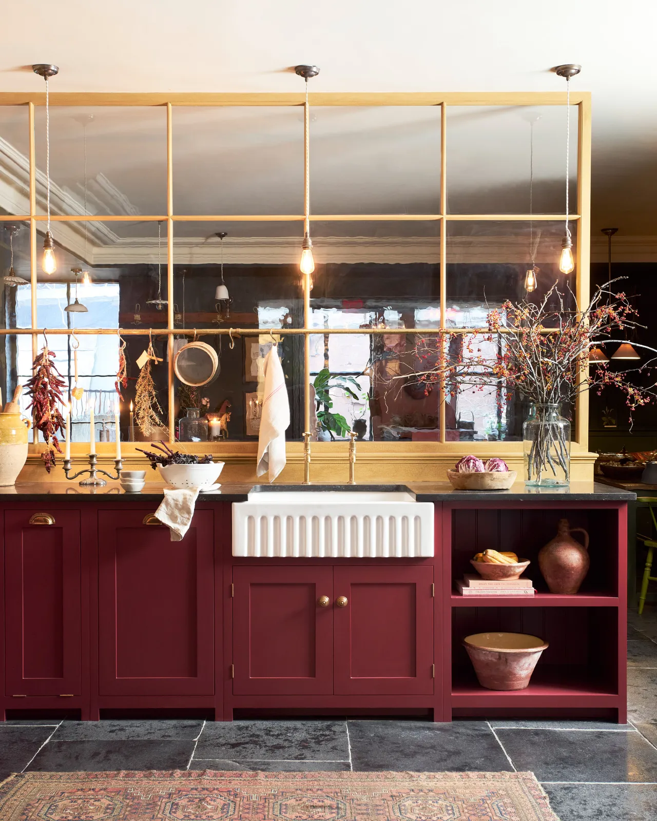 A straight-on view of a Shaker-style kitchen sink cupboard with a fluted Belfast sink, red painted cupboard doors and Victorian-inspired ornate doorknobs in a warm burnished brass finish.
