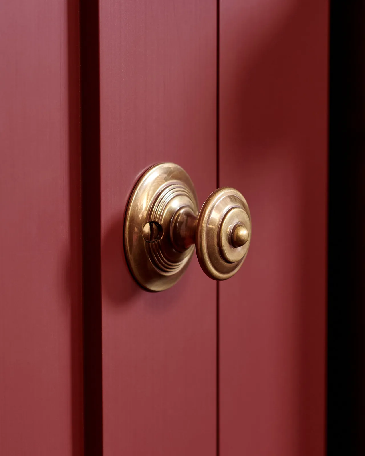 A close-up oblique view of a Victorian-inspired ornate doorknob in a warm burnished brass finish, fitted to red painted cupboard door.
