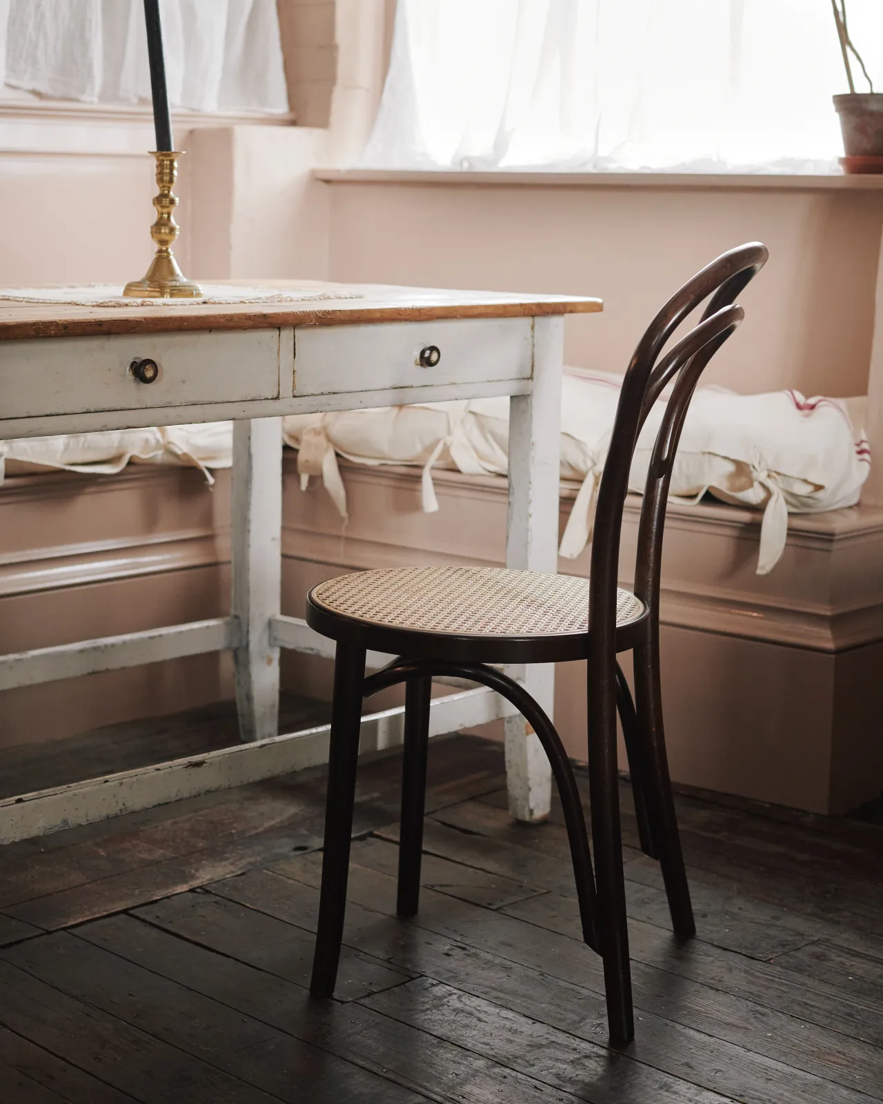 An oblique view of a café-style bentwood chair with a dark stained frame and light woven rattan seat mesh in front of a vintage pine table.