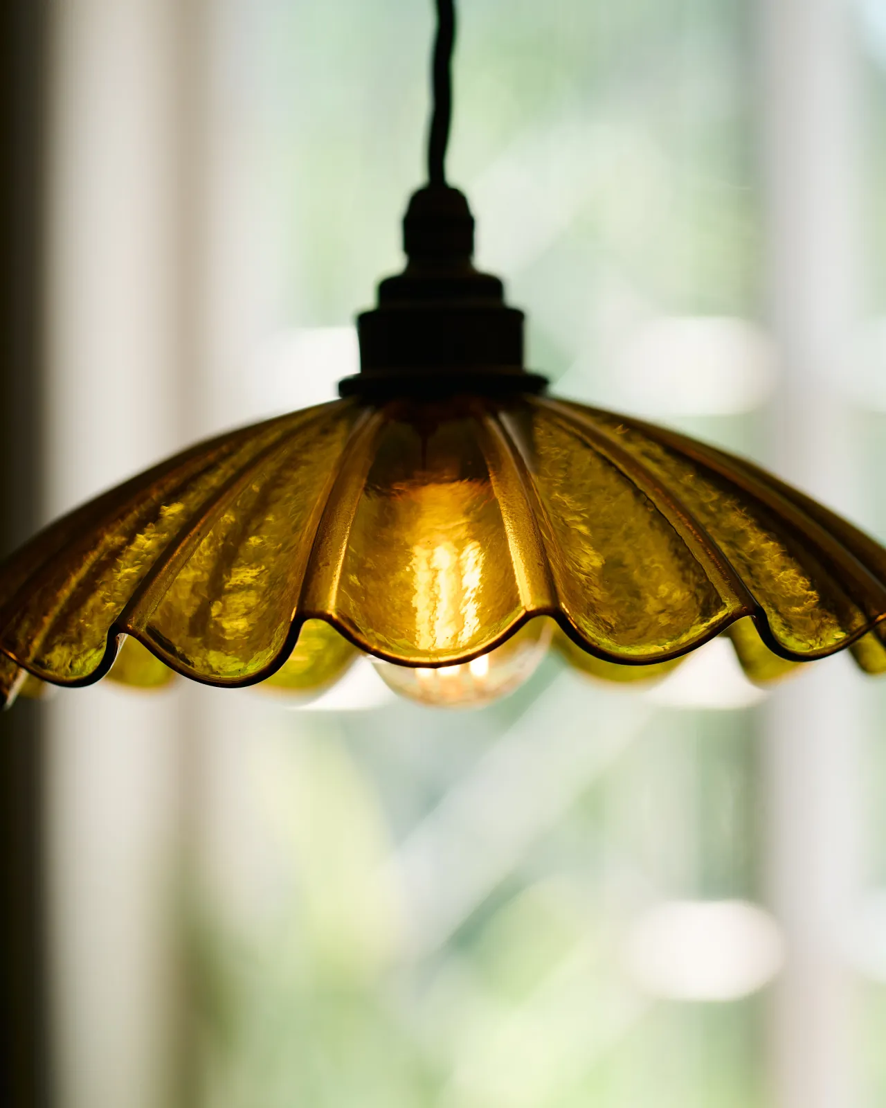 A close-up view of a small frilly chartreuse glass pendant light with brass fittings and brown twisted fabric flex cord. The light is on and shining through the tranlucent glass.