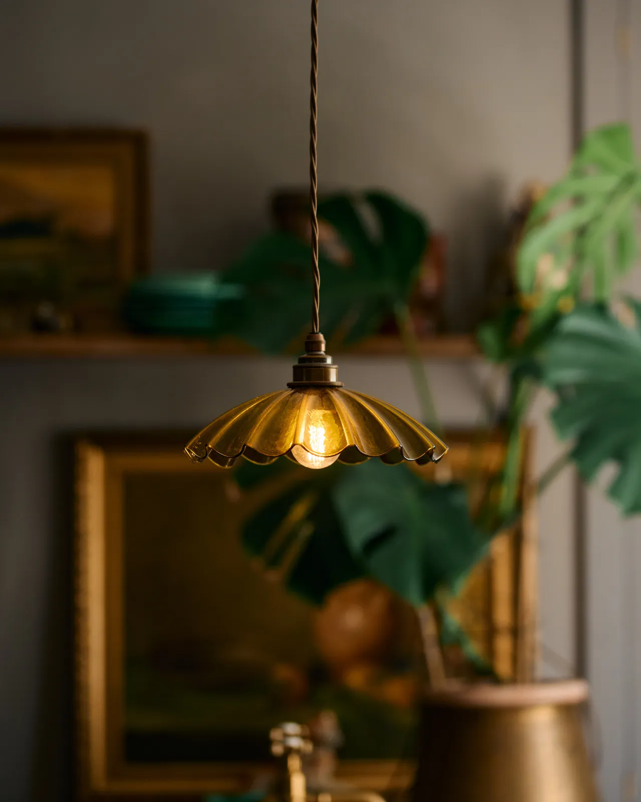 A close-up view of a small frilly chartreuse glass pendant light with brass fittings and brown twisted fabric flex cord. The light is on and shining through the tranlucent glass.