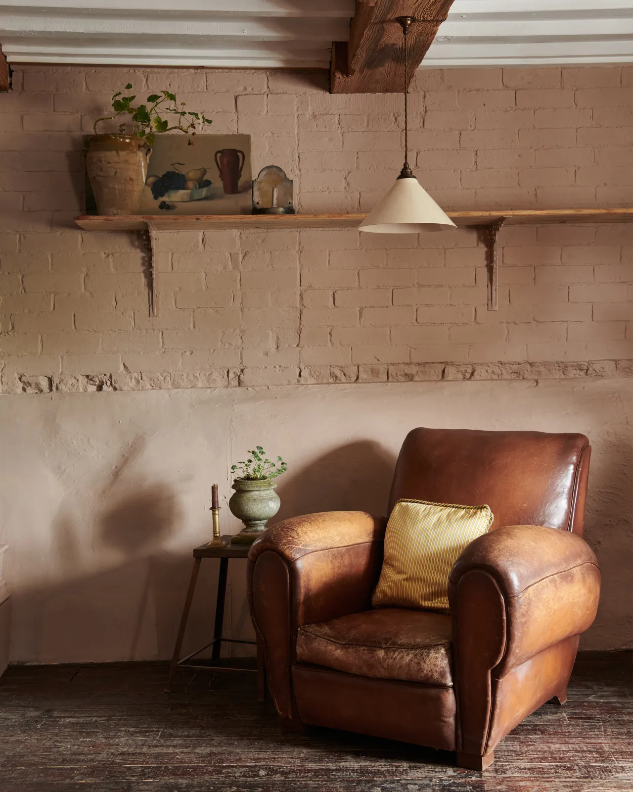 A vintage leather armchair in front of a pink brick wall and long pine shelf with Victorian-inspired ornate pink painted shelf brackets.
