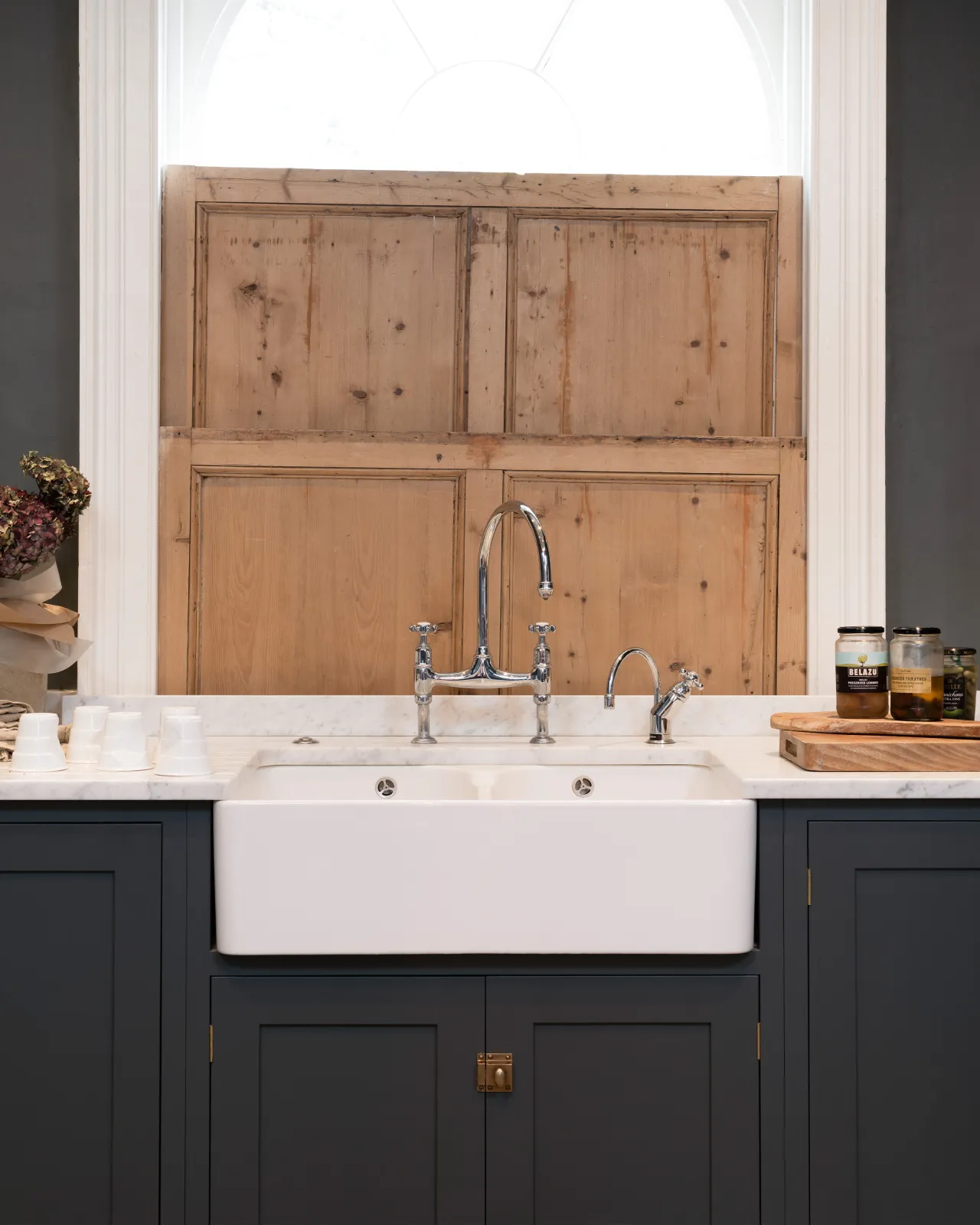 A straight-on view of a traditional-style chrome mixer tap with crosshead handles in a charcoal grey painted Shaker-style kitchen with traditional pine shutters behind.